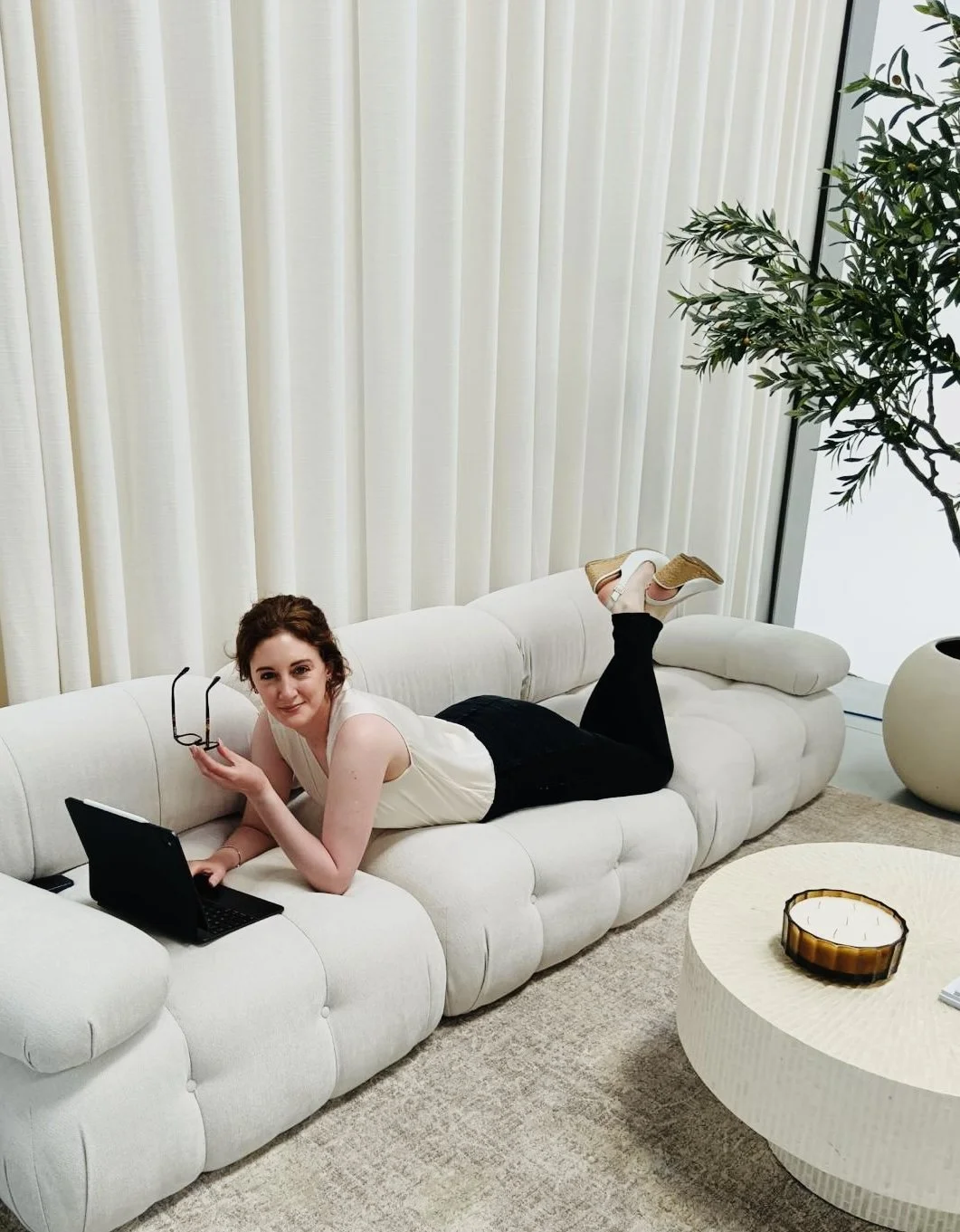 A woman lying on her stomach on a white, modern sofa, holding glasses in one hand and using a tablet with the other. She has curly brown hair, a sleeveless cream top, and black pants. The room has beige curtains, a large green plant, a round beige coffee table with a decorative bowl, and a textured beige rug.