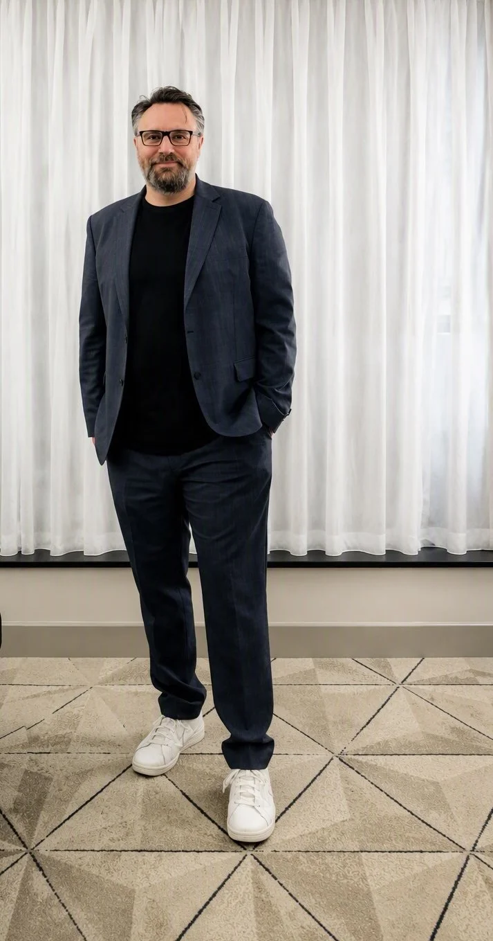 A man standing in front of white curtains, wearing glasses, a dark suit jacket, black shirt, dark pants, and white sneakers.
