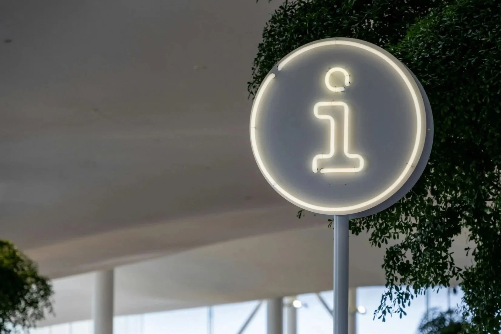 Neon sign displaying the letter 'i' for information, mounted outdoors near trees.