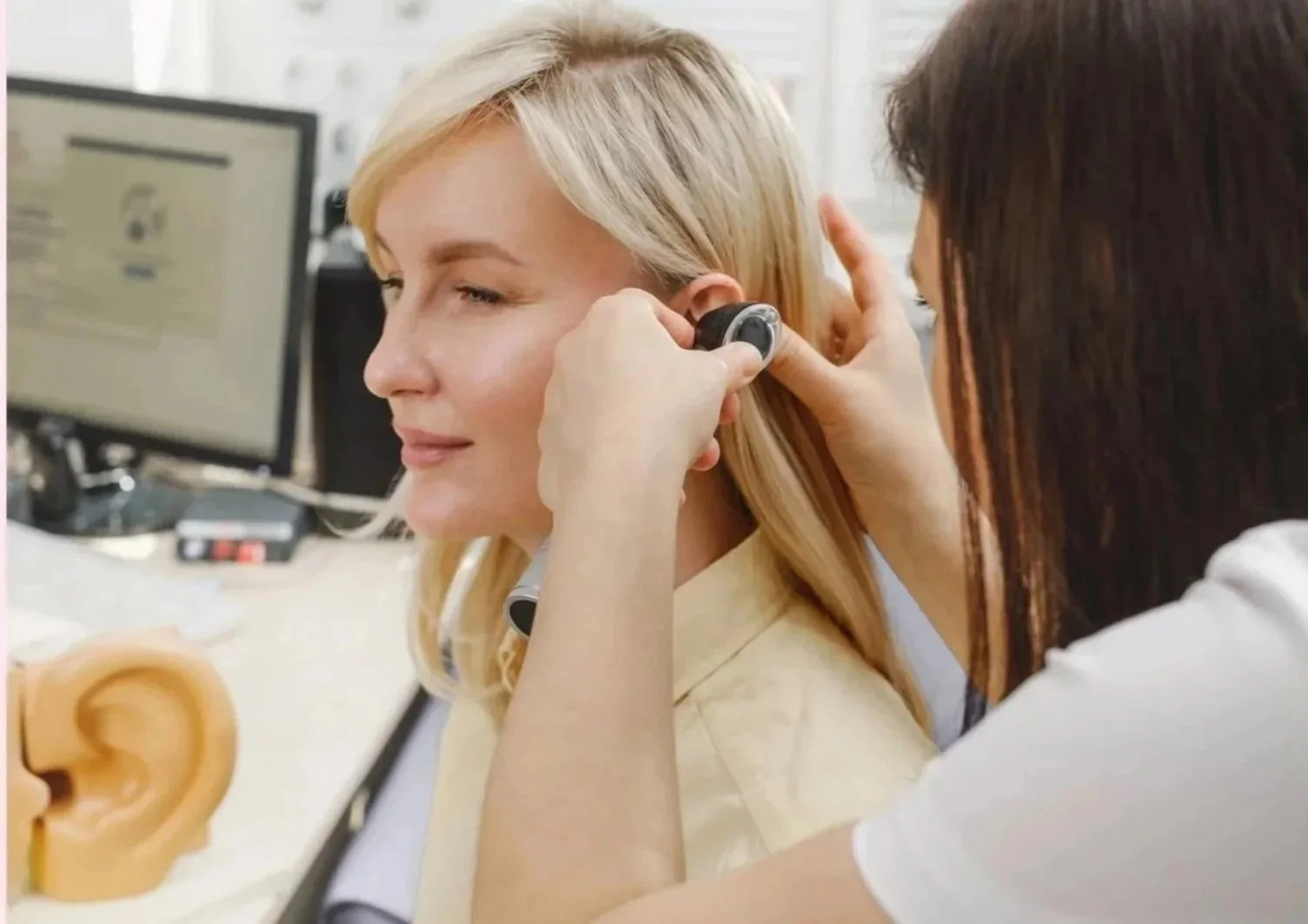 A woman with blonde hair has an ear exam being performed by a healthcare professional in an office setting, with a computer monitor in the background.