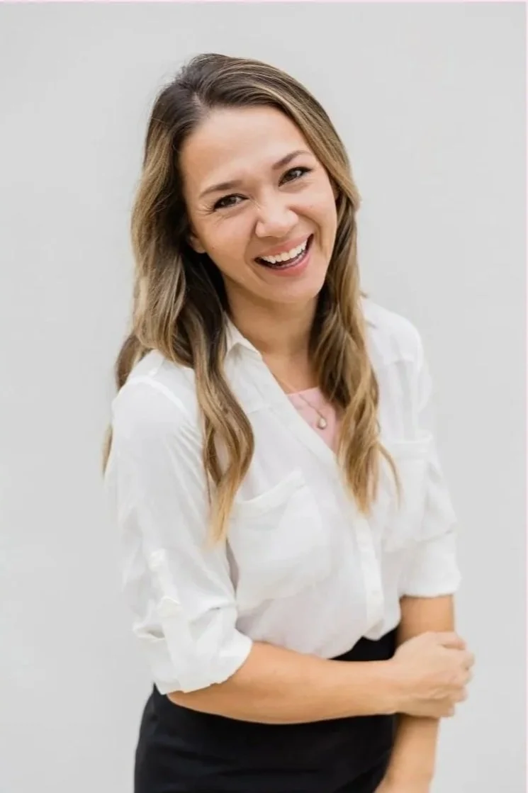 A woman with long, wavy brown hair smiling and wearing a white blouse.