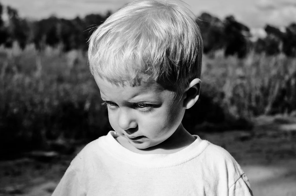 Black and white photo of a young boy looking down, outdoors in a natural setting.