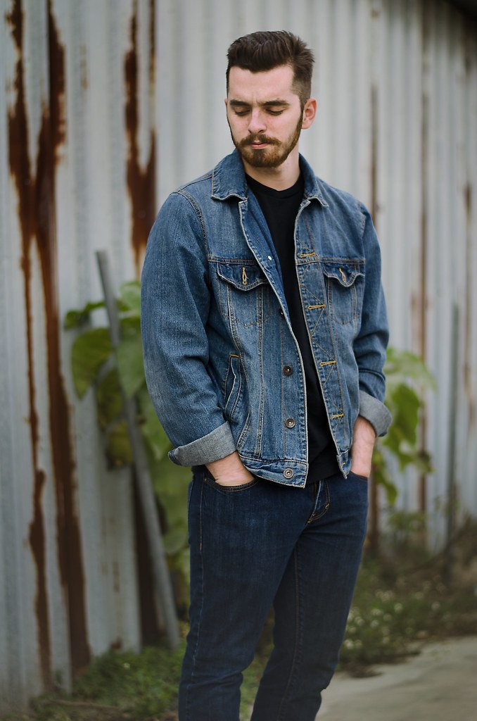 A man with a beard and short dark hair, wearing a denim jacket and black pants, stands outdoors near a rusty corrugated metal wall with green plants in the background, looking down with his hands in his pockets.