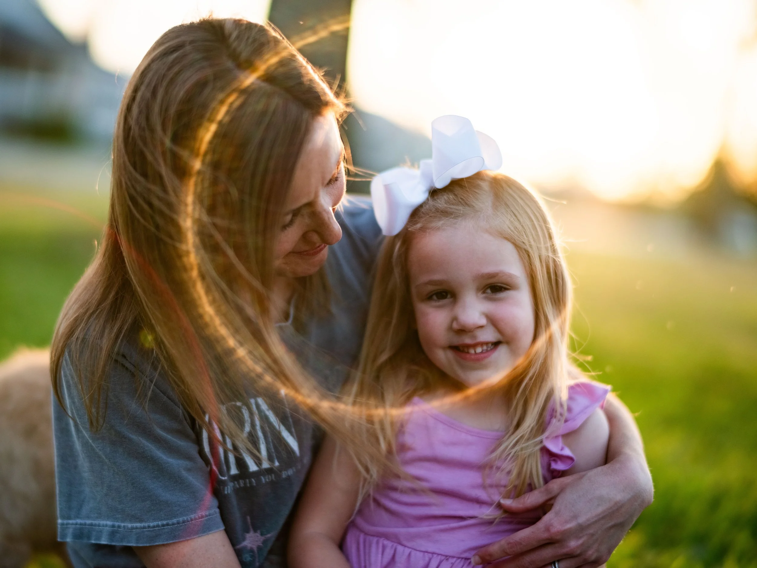 A woman with long hair and a young girl with a large white bow in her hair are outdoors, embracing each other and smiling as the sun sets behind them.