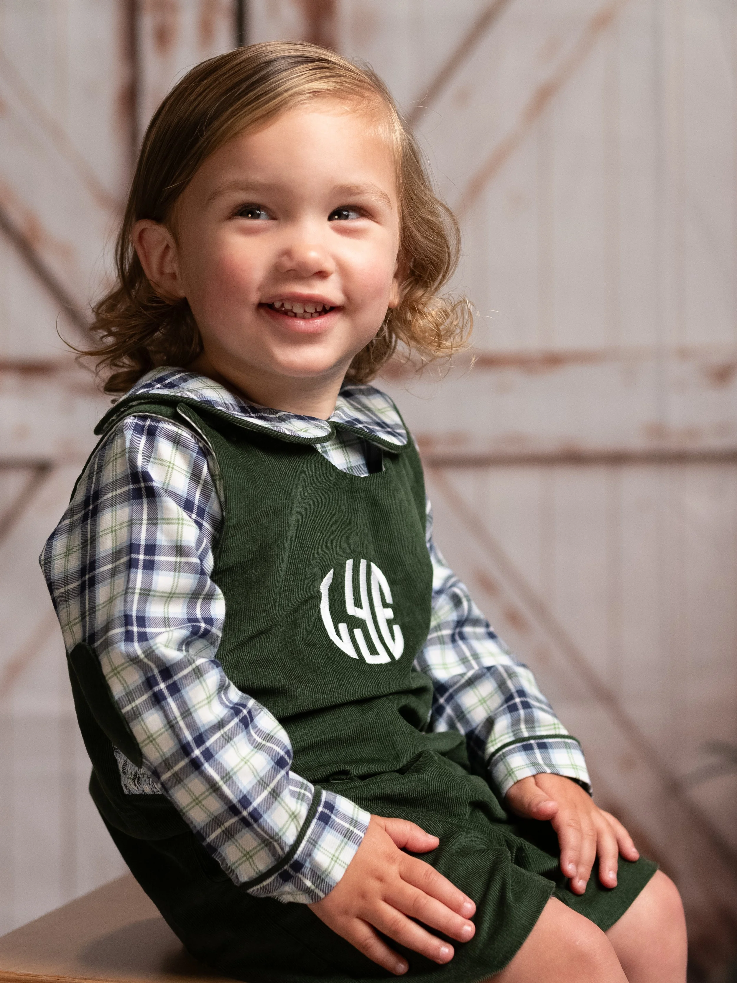 A young girl with long, light brown hair sitting on a chair. She is smiling, showing her missing front teeth. She is wearing a blue plaid shirt under a dark green apron with a monogram. The background is a wooden wall.