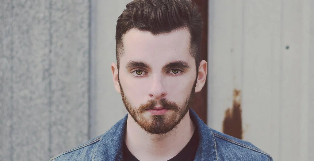 A young man with dark hair, a beard, and green eyes, wearing a denim jacket, standing in front of a weathered wooden wall.