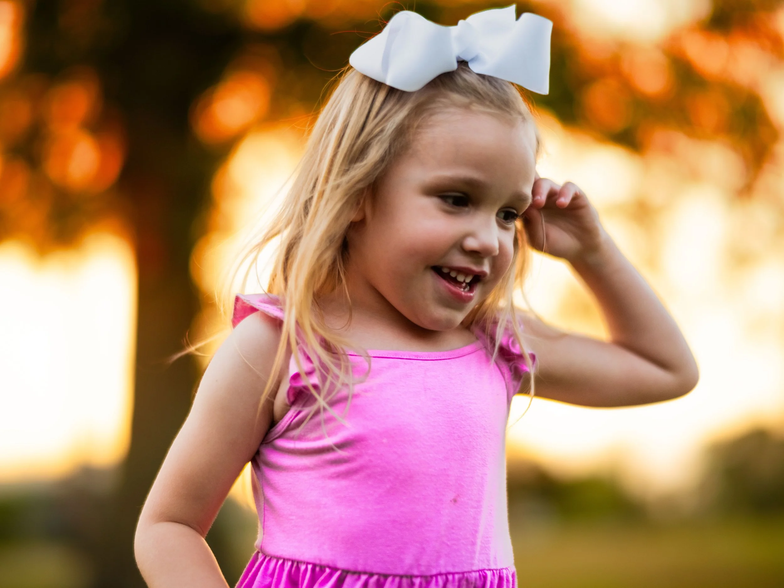 A young girl with long blonde hair wearing a pink sleeveless top and a large white bow on her head, smiling outdoors during sunset with blurred orange and yellow background.