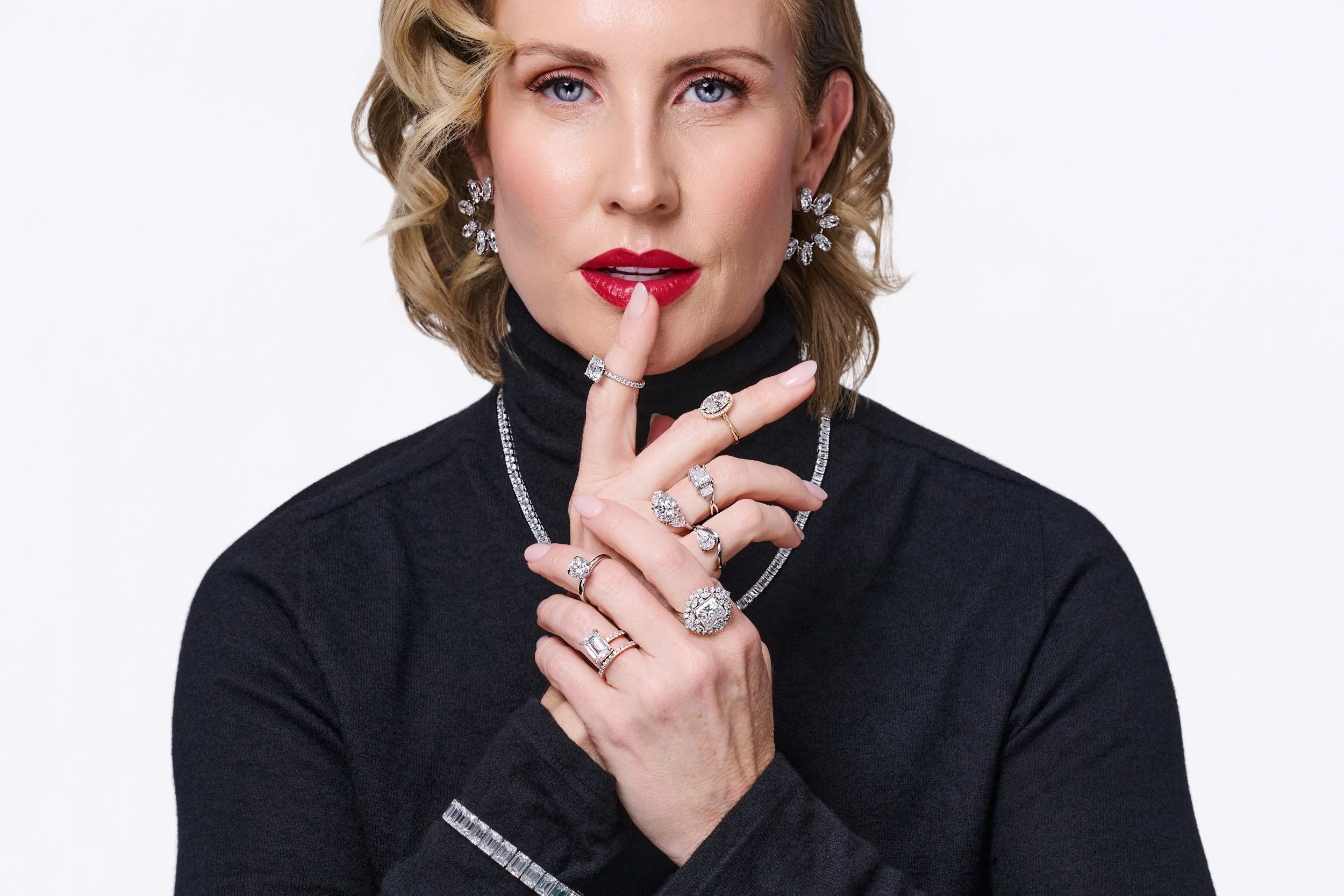 Close-up of a woman with blonde hair, wearing a black top, red lipstick, and jewelry including earrings and multiple rings, holding her finger to her lips against a plain background.