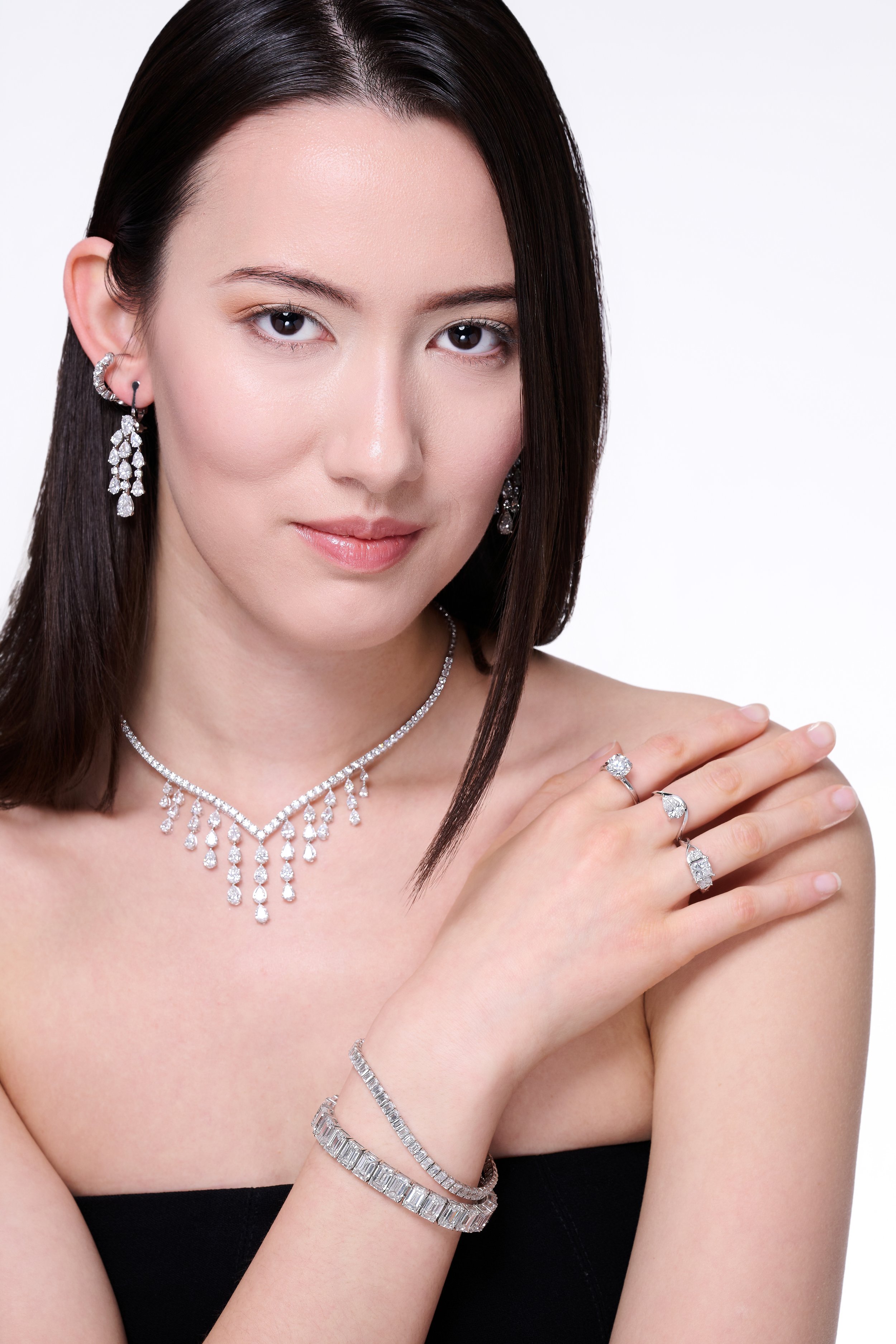 A woman with dark brown hair and fair skin wearing diamond jewelry including earrings, a necklace, rings, and bracelets, posing with her hand on her shoulder against a white background.