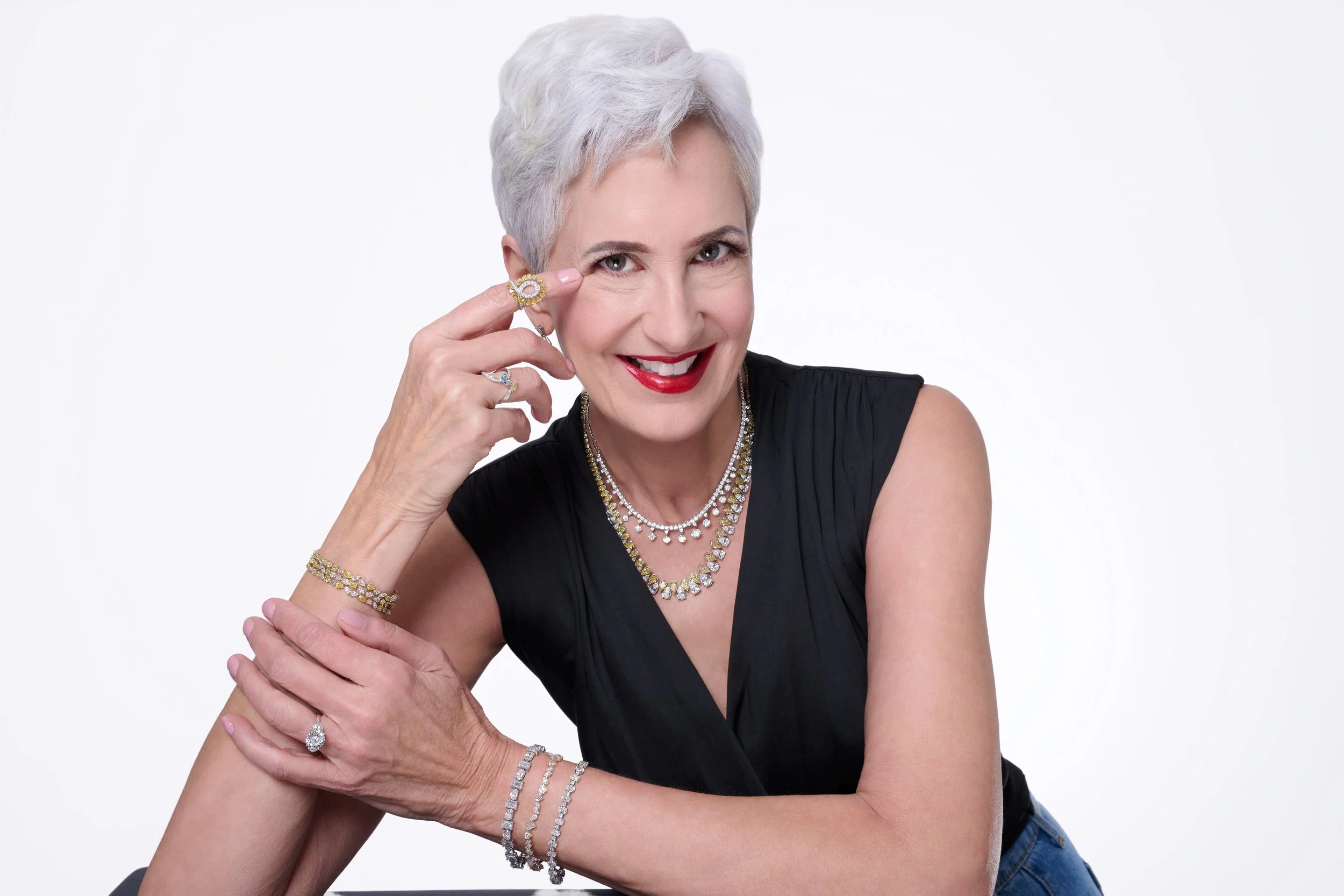 An older woman with short, white hair, smiling, wearing a black top, and sitting close to another person. She is posing with her hand near her face, wearing multiple pieces of jewelry including rings, bracelets, a necklace, and a ring on her finger.