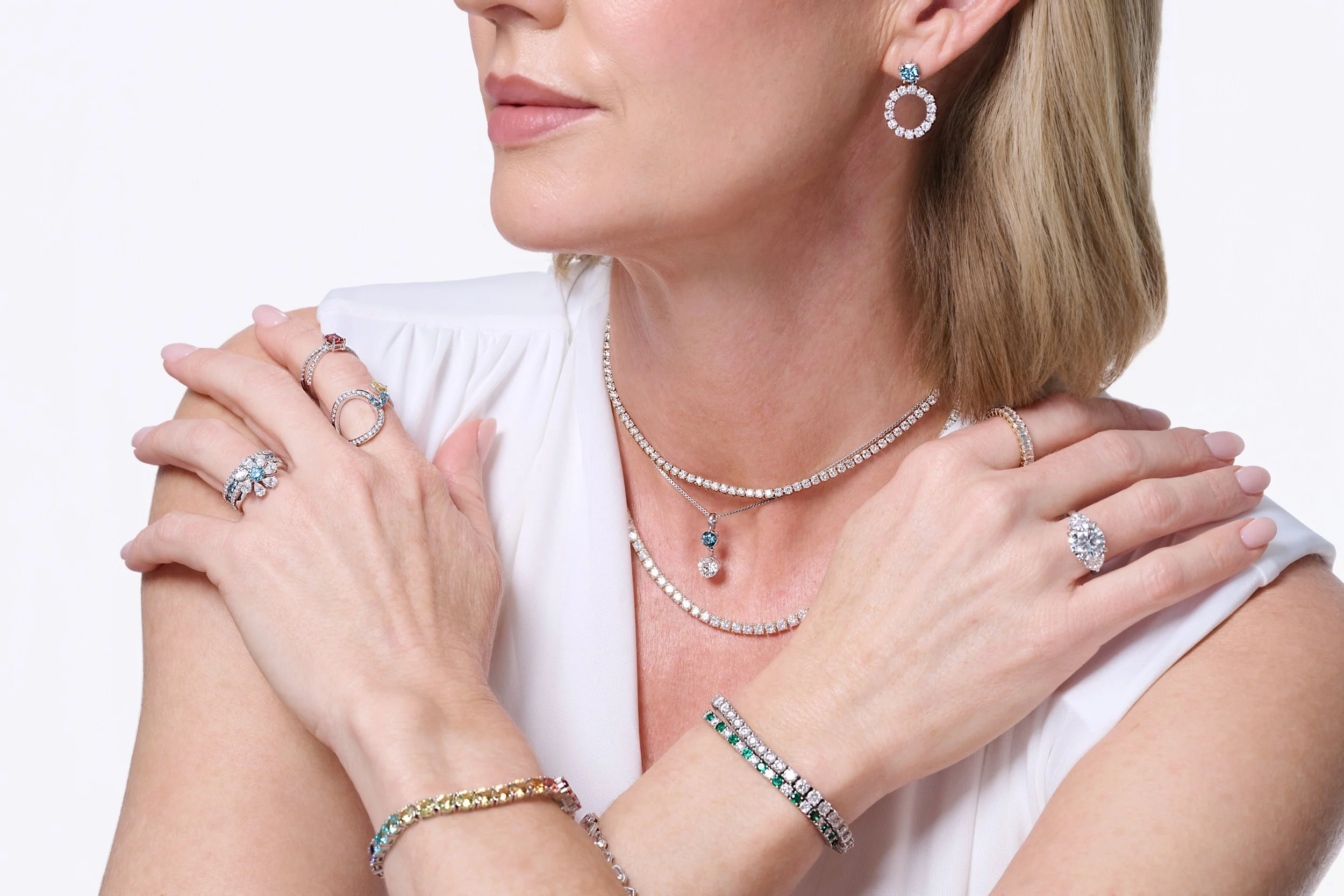 Close-up of a woman wearing multiple pieces of diamond jewelry, including earrings, necklaces, rings, and bracelets, with a white top against a plain white background.