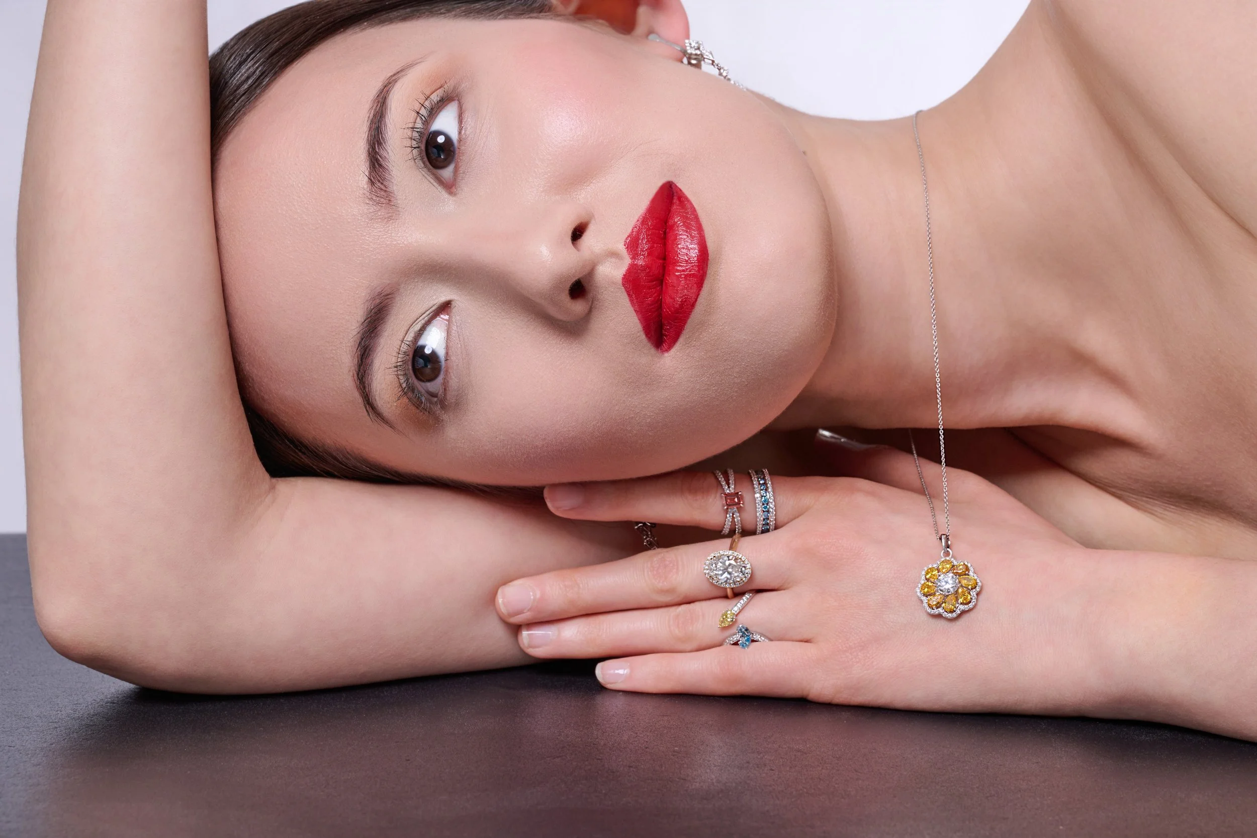 A woman with fair skin and dark brown hair lying on a black surface, resting her head on her arm, wearing multiple rings and jewelry, with bright red lipstick, eye makeup, and earrings.