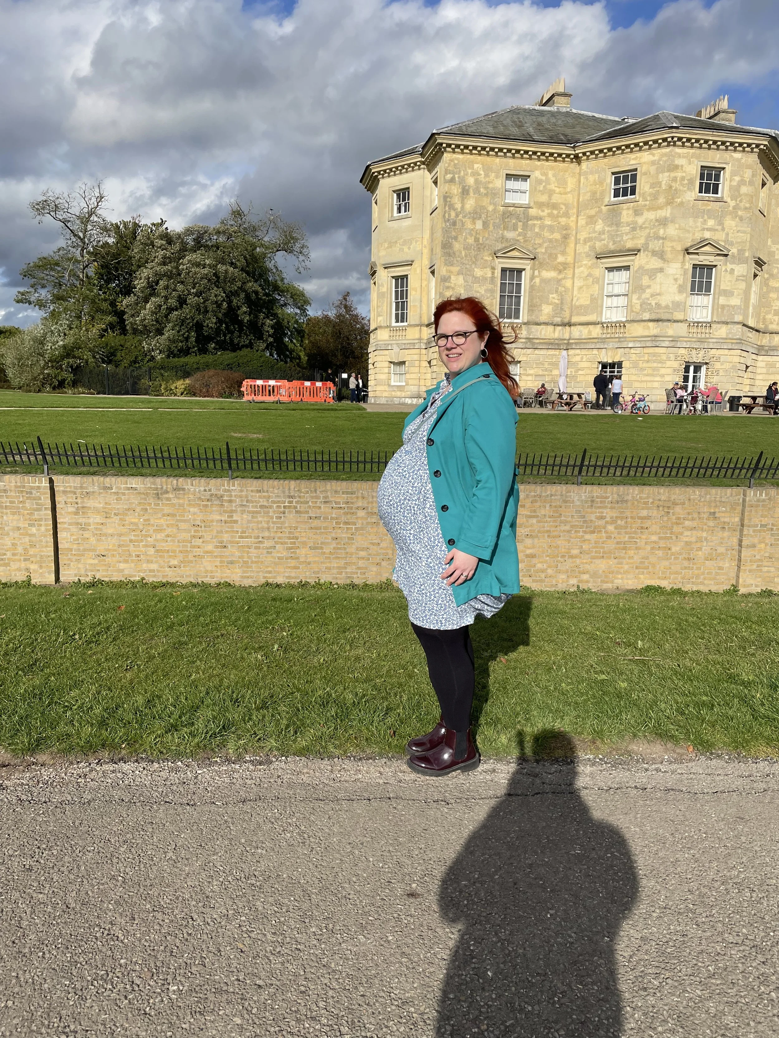 A pregnant woman with red hair and glasses standing outside on a sunny day, wearing a light blue jacket, a patterned dress, black leggings, and maroon boots. Behind her is an old stone building, a grassy area, some trees, and a black metal fence. There are people, bicycles, and outdoor seating in the background.