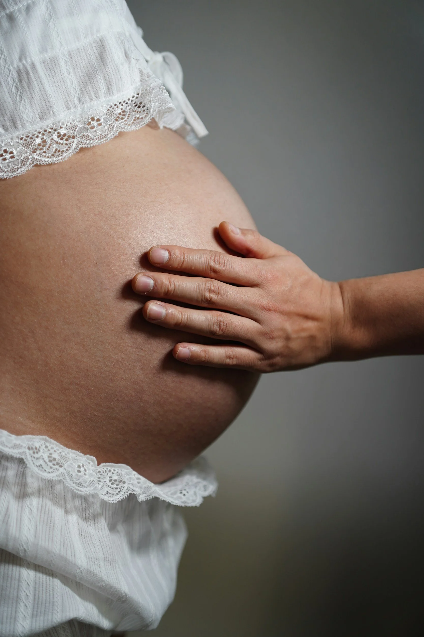 A pregnant woman wearing a white lace dress shows her belly, with her hand resting on it against a neutral gray background.
