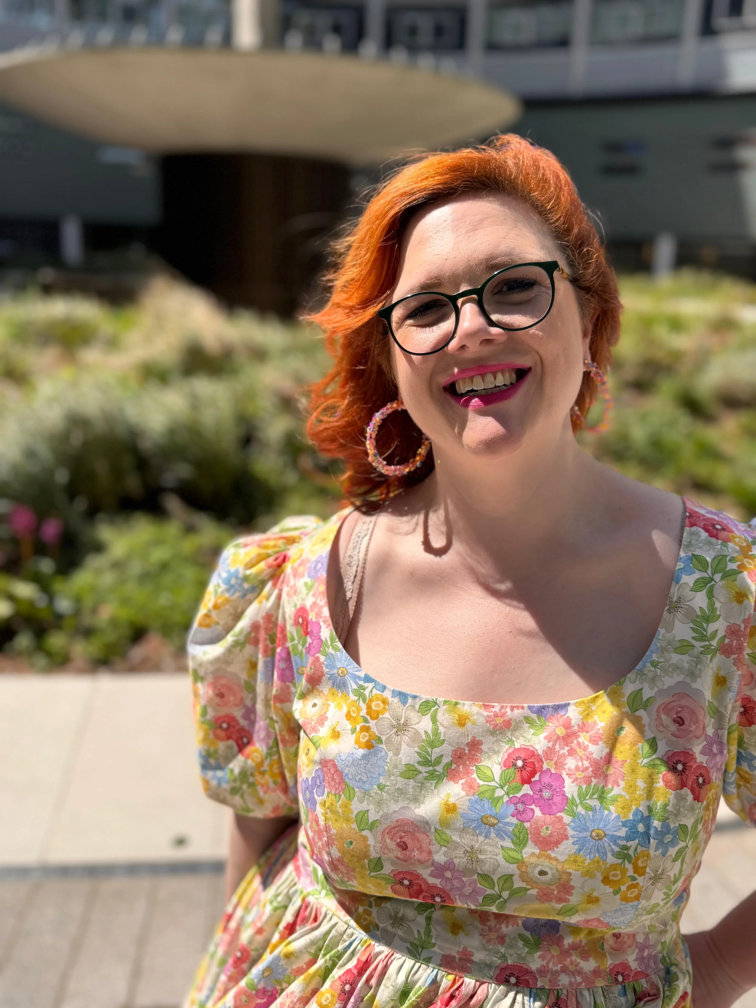 A woman with red hair, glasses, and earrings smiling outdoors in front of a building with a garden in the background, wearing a colorful floral dress.