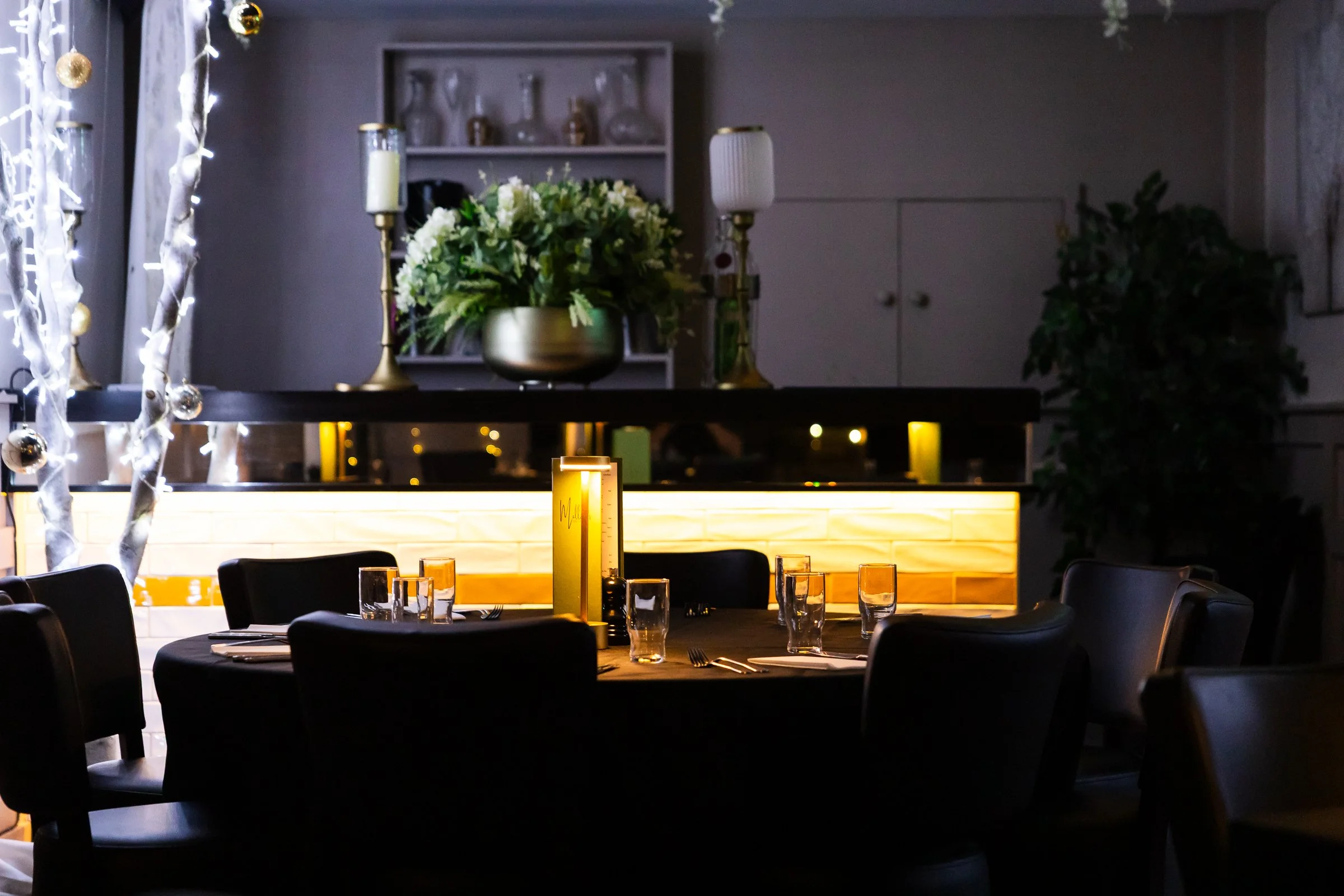 A decorated indoor dining area with tables set with glasses, napkins, and cutlery, illuminated by warm lighting, with a floral centerpiece, candlesticks, and festive ornaments in the background.