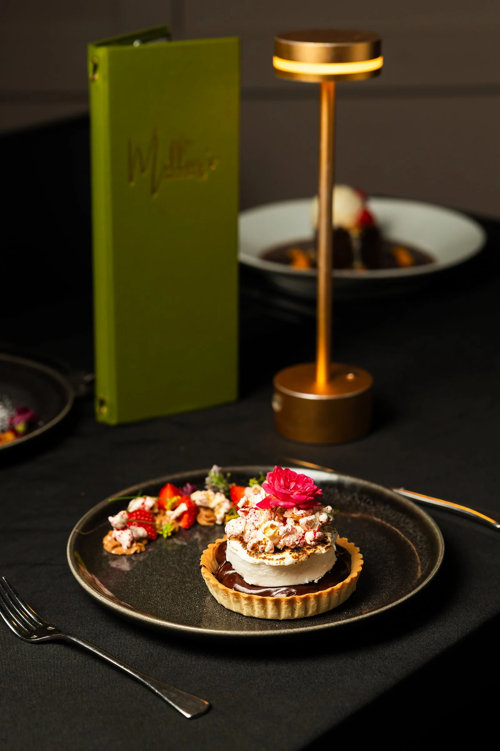 A gourmet dessert tart with whipped cream, popcorn, and edible flowers, served on a black plate with a fork, with a dark background featuring a green menu and a decorative lamp.