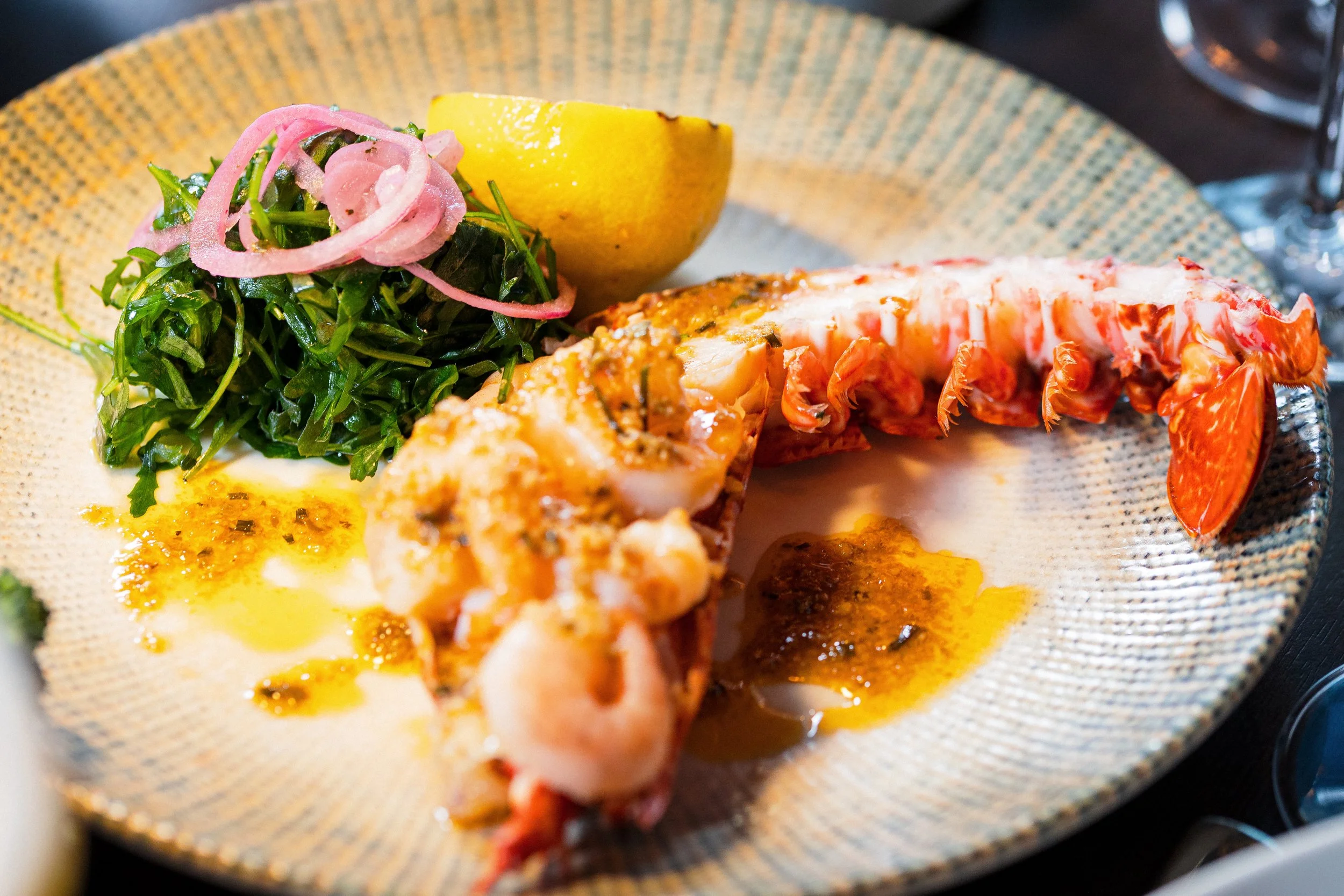 Lobster tail with melted butter, a lemon wedge, and a greens salad with pickled onions on a decorative plate.