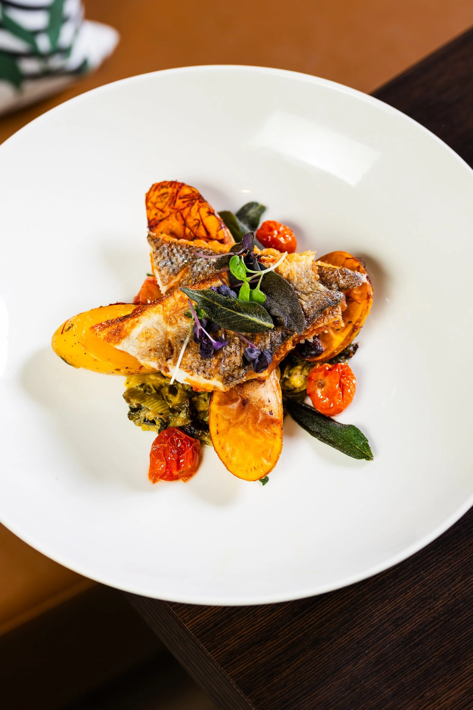 A plated dish featuring a cooked fish fillet topped with microgreens, surrounded by roasted cherry tomatoes, with sliced yellow bell peppers and sautéed greens underneath, on a white round plate.