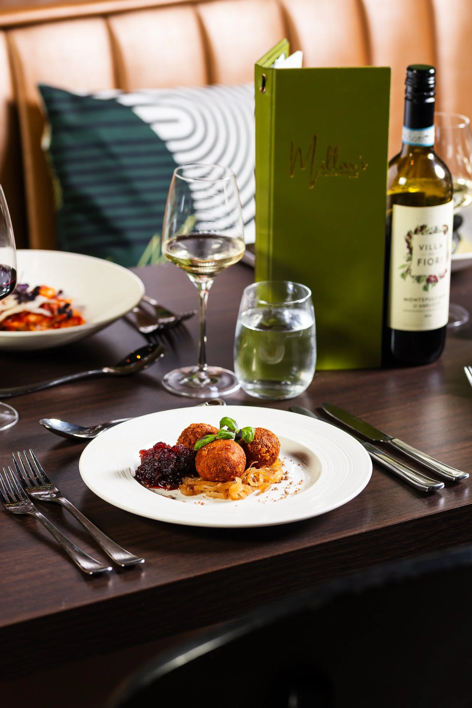 A dinner table with a white plate of fried appetizers garnished with basil leaves, served with a side of sauce, along with a glass of white wine, a glass of water, a green menu labeled 'Villa Fiori', a bottle of white wine, and cutlery, in a dining s