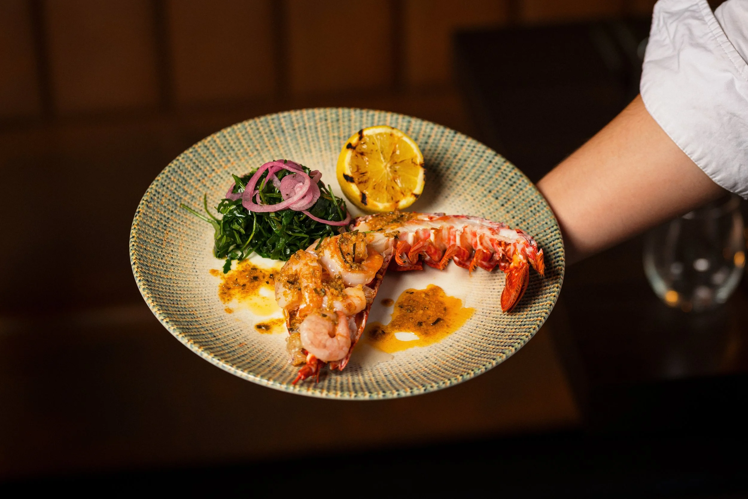 A plate of cooked lobster tail with lemon wedge, green salad with pickled red onions, and a drizzle of butter or sauce on the lobster.