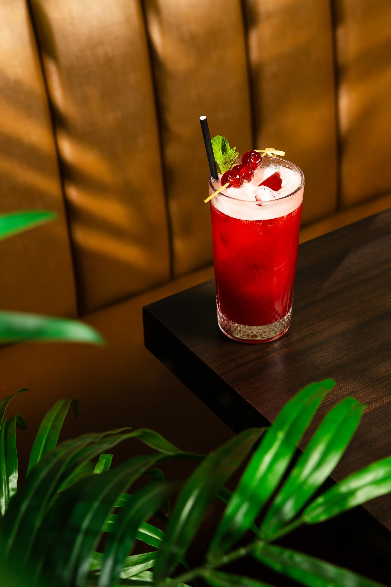 A tall glass of red cocktail with ice, garnished with red berries, a sprig of mint, and a small decorative skewer, placed on a dark wooden table with green leafy plants in the foreground and a brown upholstered wall in the background.