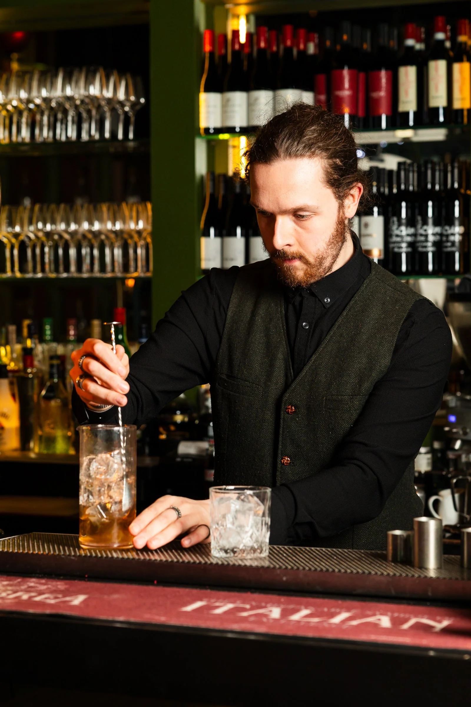 A bartender with long hair and a beard prepares a drink behind a bar, with shelves of wine bottles and glasses in the background. There are also glasses and ice on the counter.