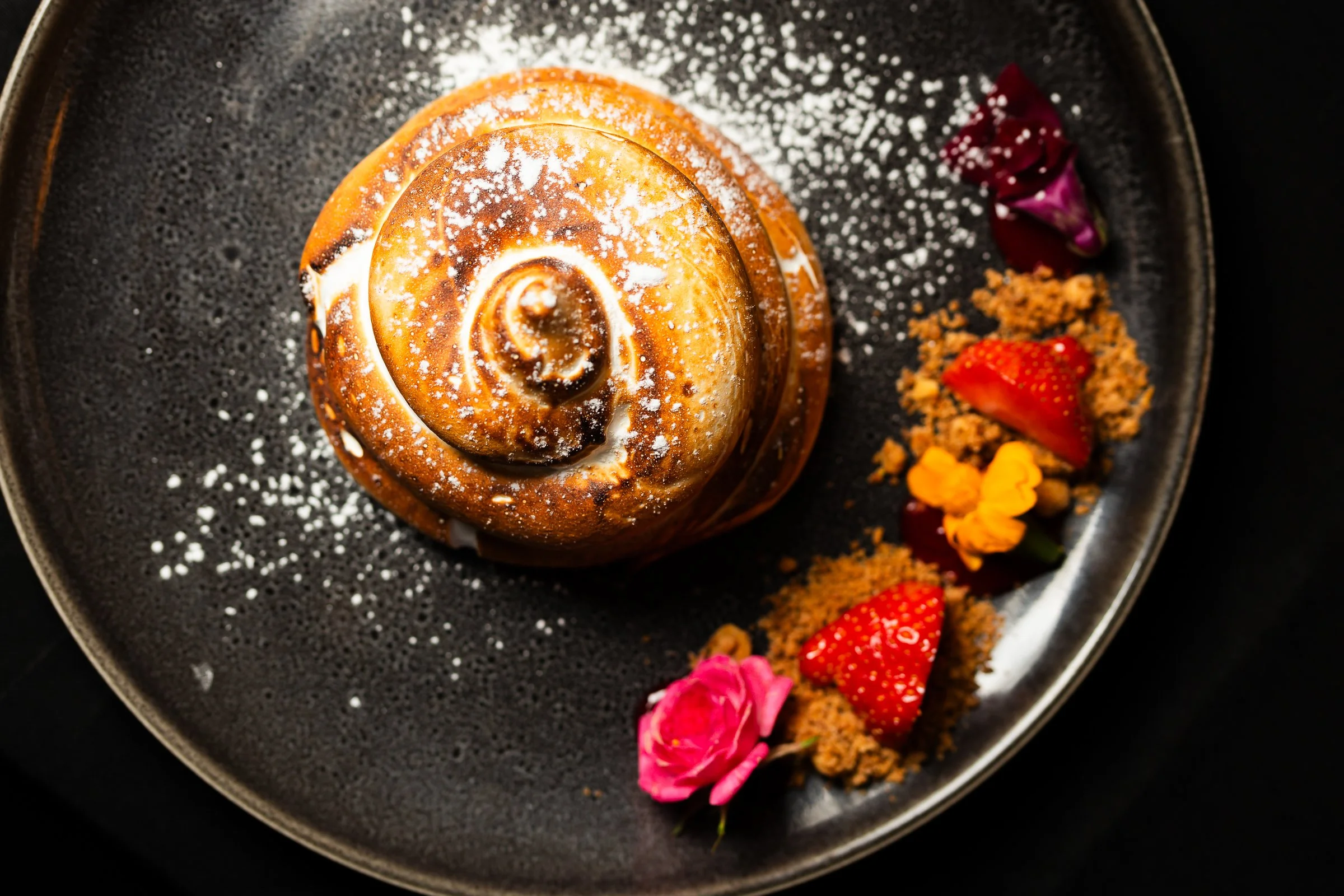 A cinnamon roll dusted with powdered sugar on a black plate, garnished with strawberries, edible flowers, and crumbled topping.