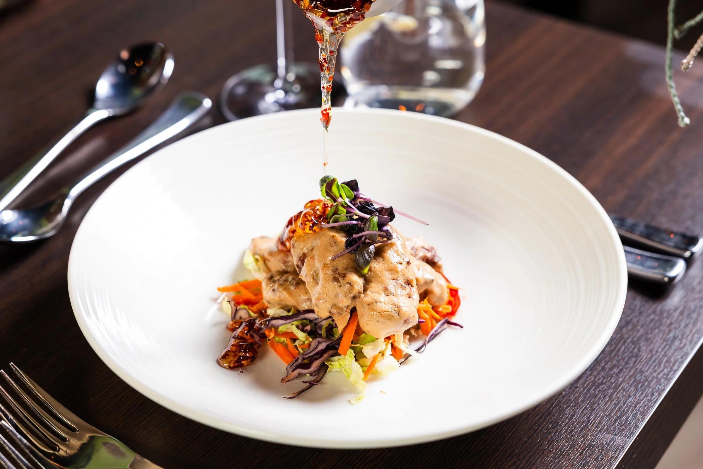 A plated dish of chicken topped with microgreens and red peppers, served over shredded carrots and cabbage, with a drizzle of sauce.