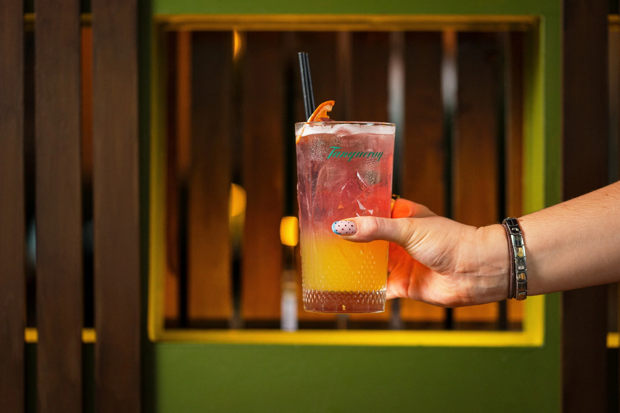 A tall glass of a layered beverage with orange slice and black straws being held by a person with painted nails, a wristwatch, and a bracelet, against a background of wooden slats and brightly colored accents.
