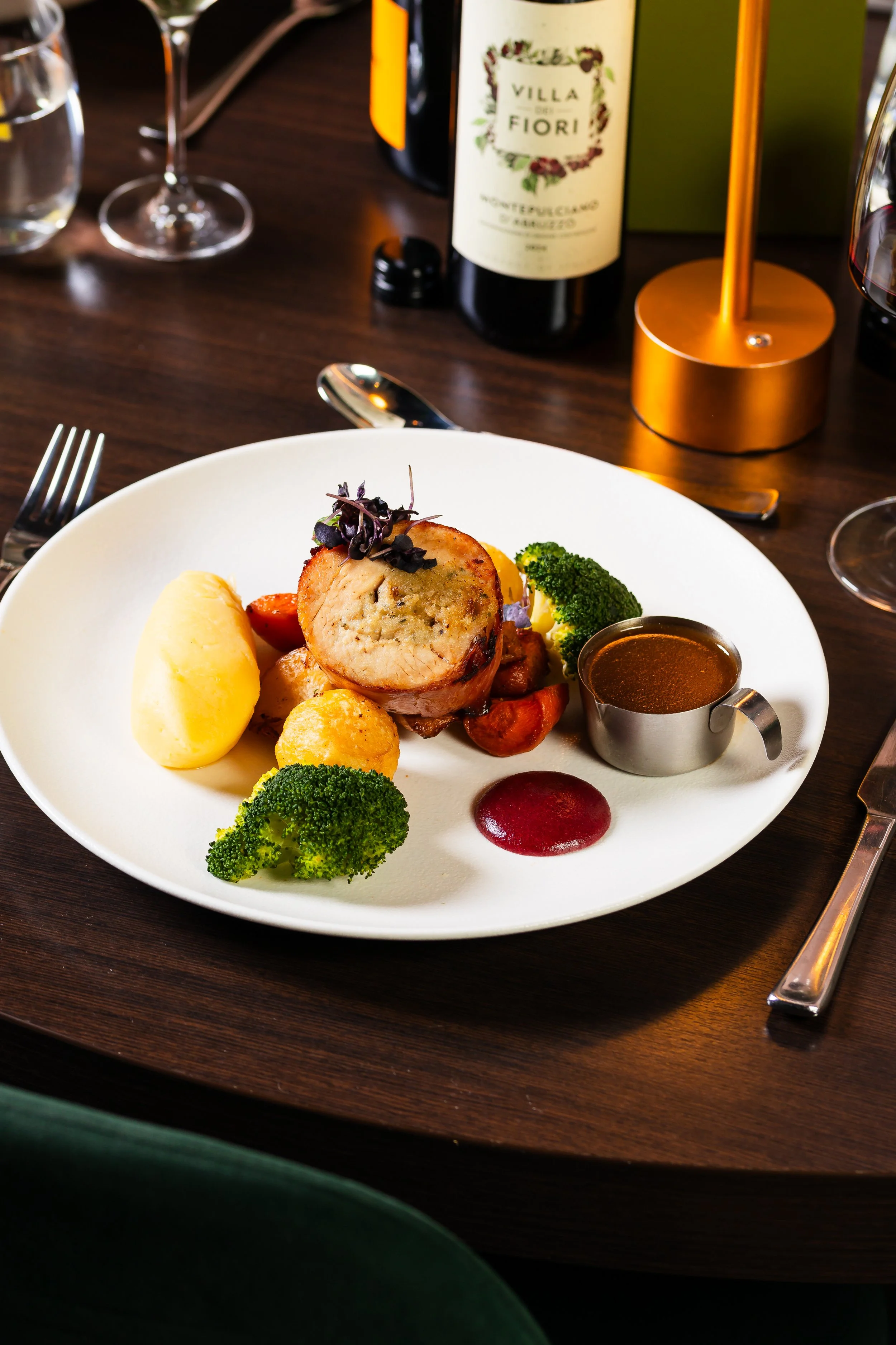 A white plate with a meatloaf slice topped with microgreens, accompanied by mashed potatoes, broccoli, cherry tomatoes, and a small cup of brown gravy. There are wine glasses and bottles in the background on a dark wooden table.