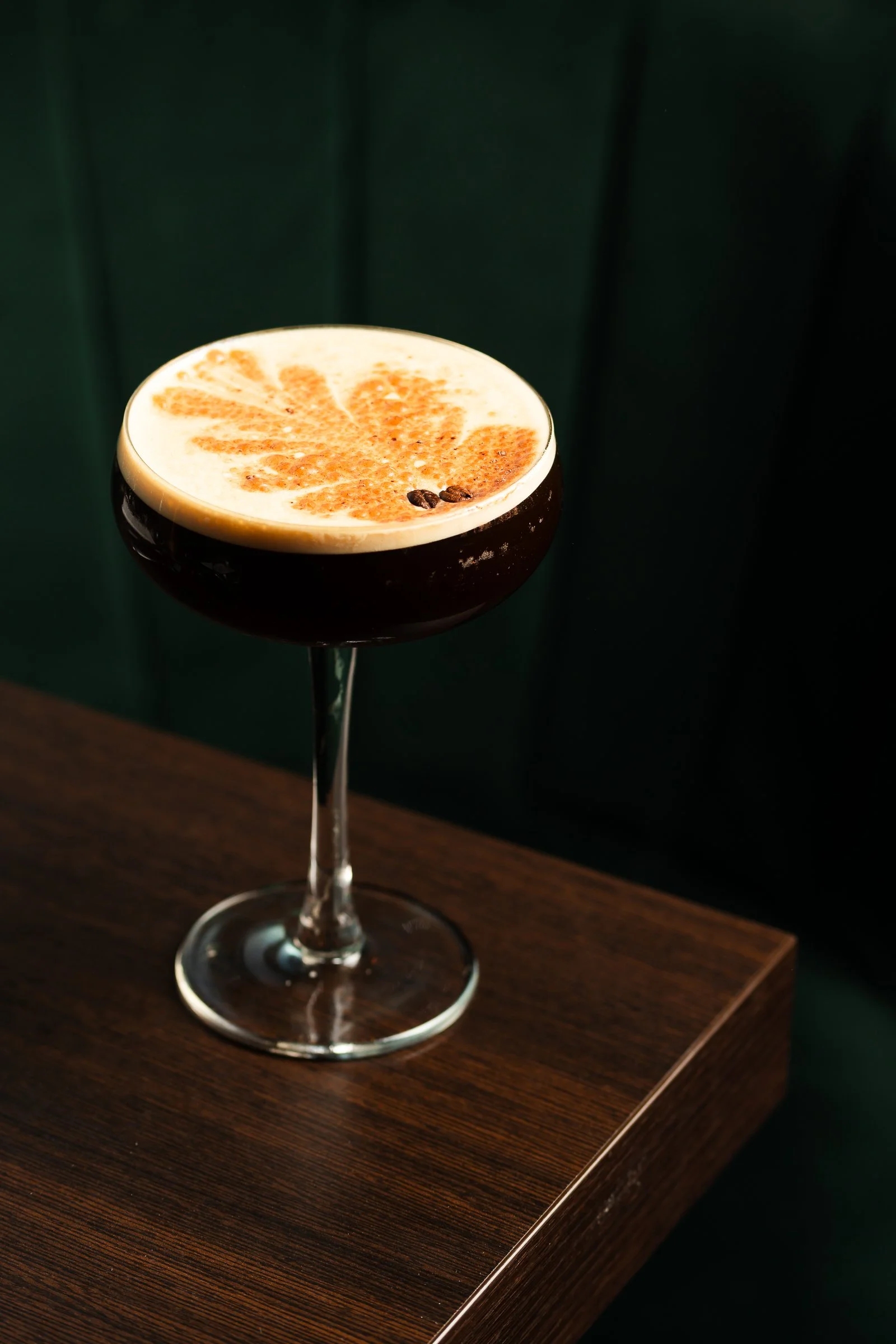 A dark cocktail glass filled with a creamy beverage topped with orange zest and coffee beans, placed on a wooden surface against a dark green background.