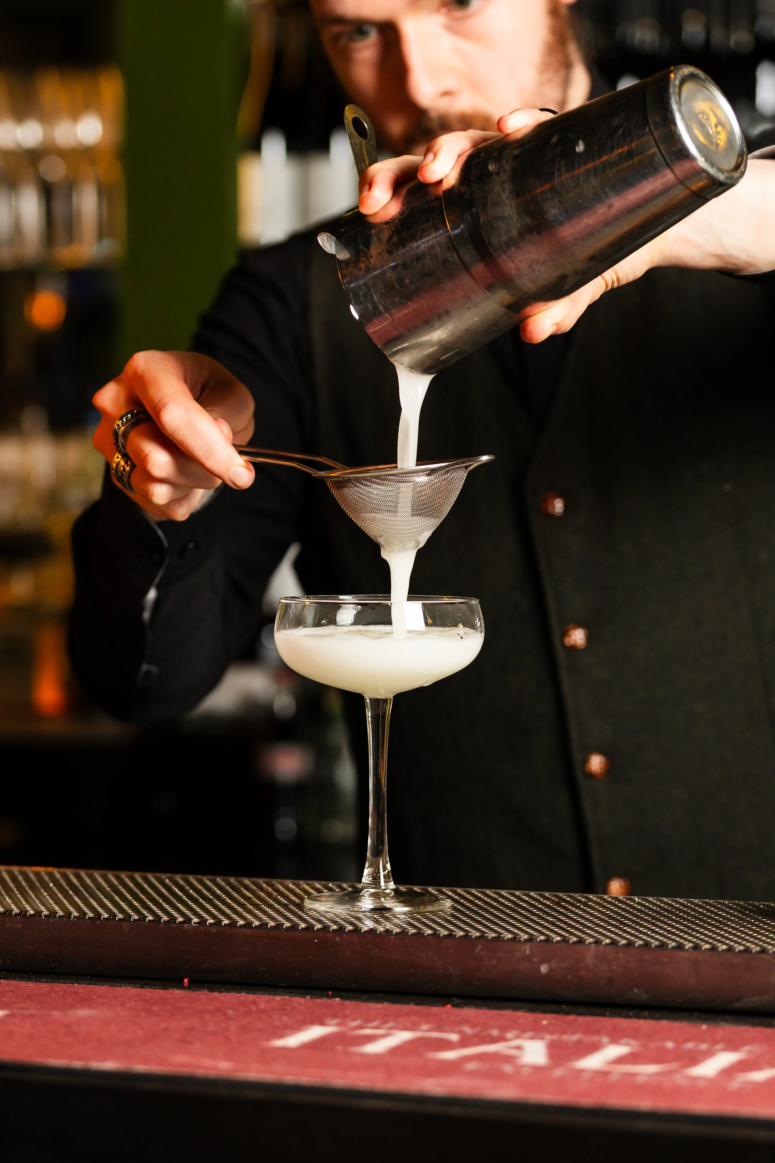 A bartender pours a white cocktail through a strainer into a coupe glass at a bar.