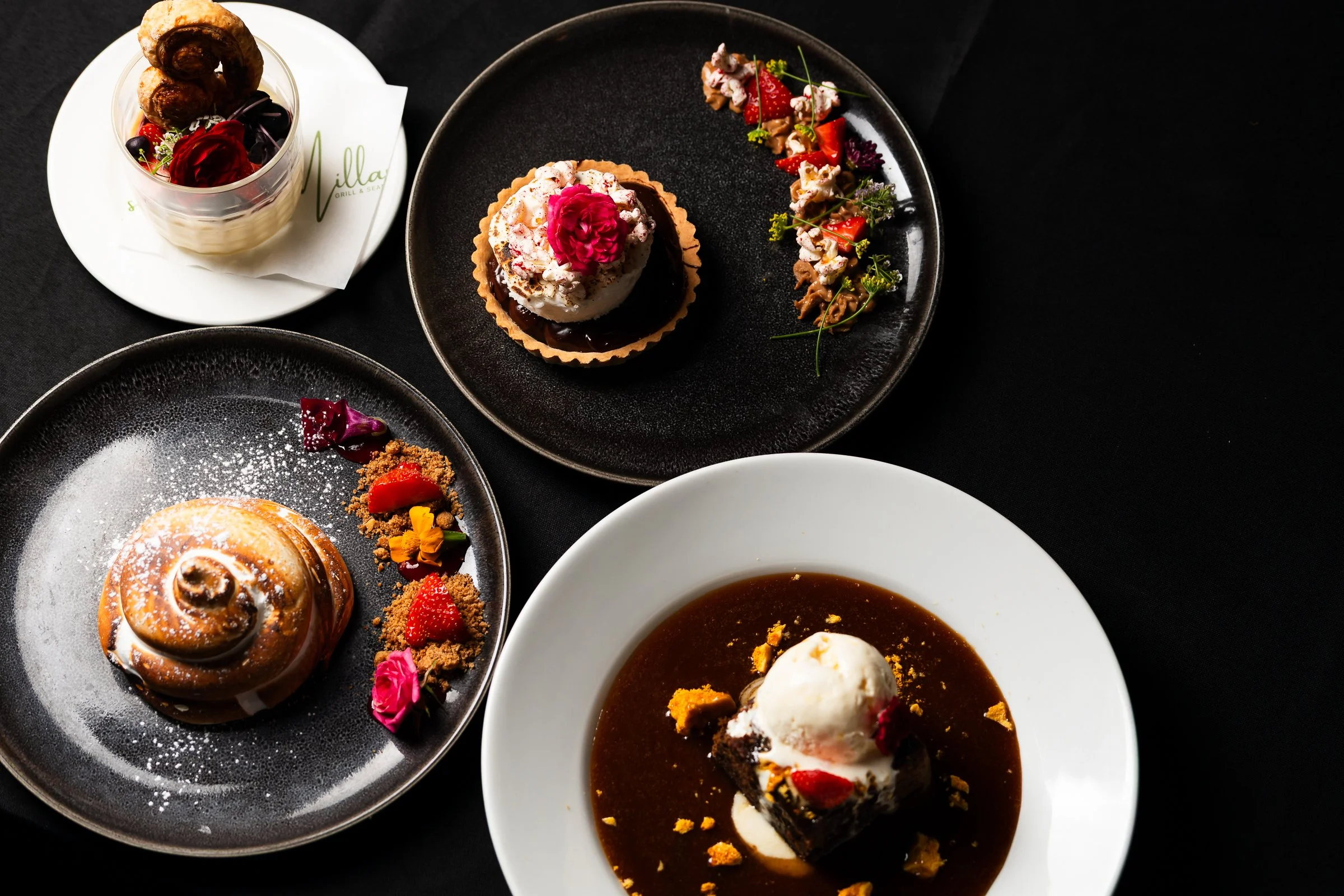 Assorted desserts on black and white plates, including a pie topped with whipped cream and a pink flower, a chocolate cake with fruit and flowers, and a cinnamon roll with powdered sugar, garnished with berries and edible flowers.