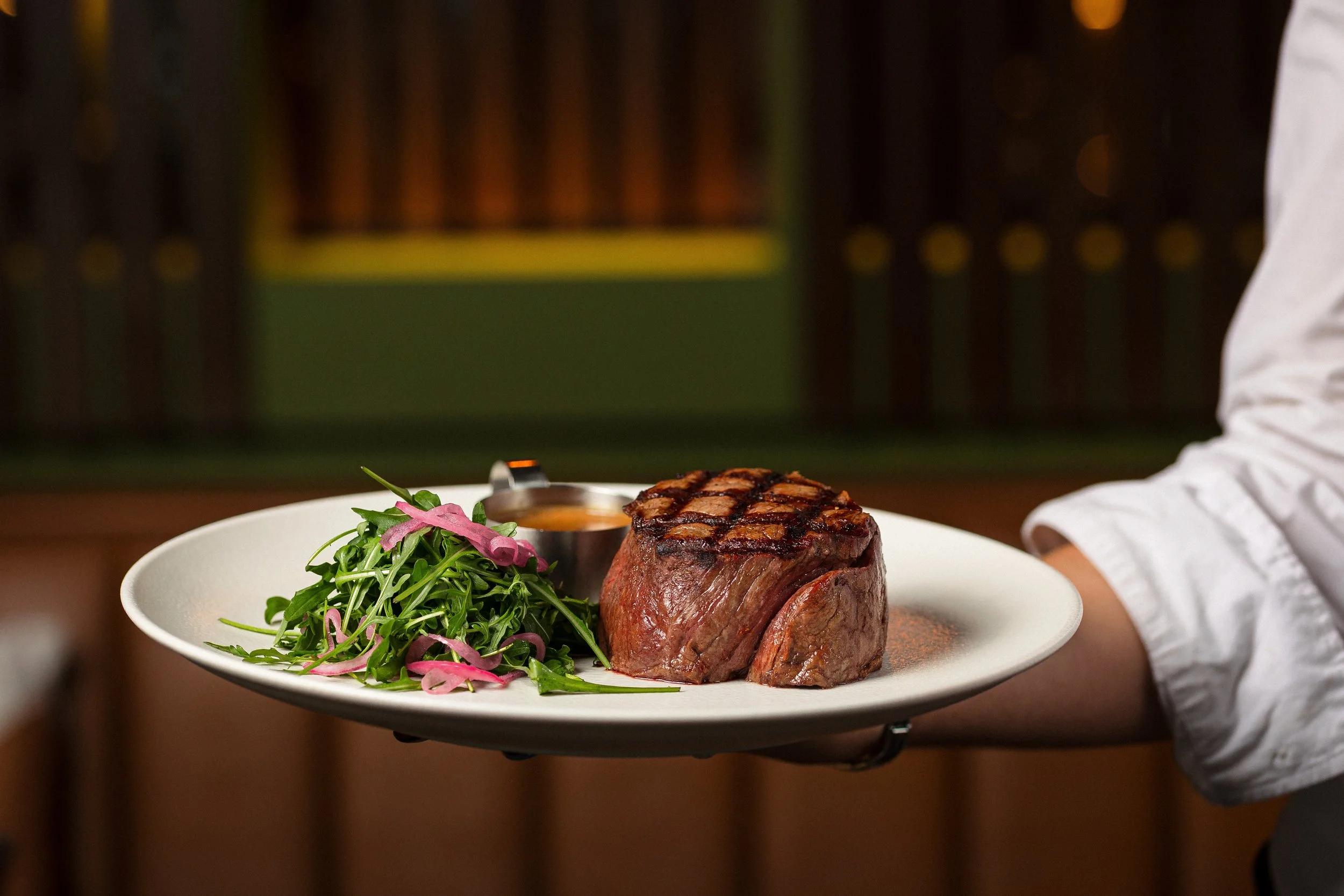 A chef holding a white plate with a grilled filet mignon, a side of mixed greens with pink pickled onions, and a small container of sauce, in a restaurant setting.