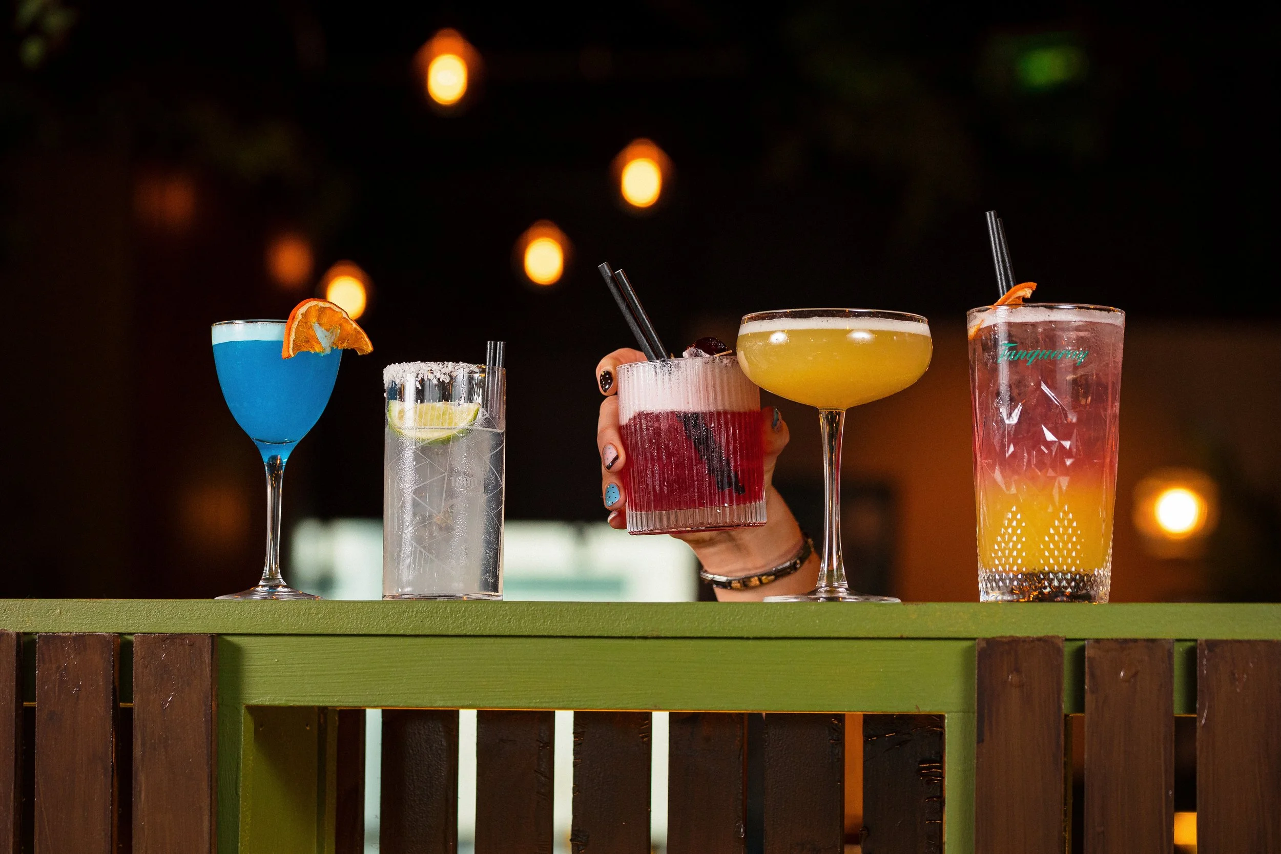 Four colorful cocktails on a bar counter with a dark background, including a blue drink with orange garnish, a clear drink with lime, a pinkish-red drink, and a yellow drink.