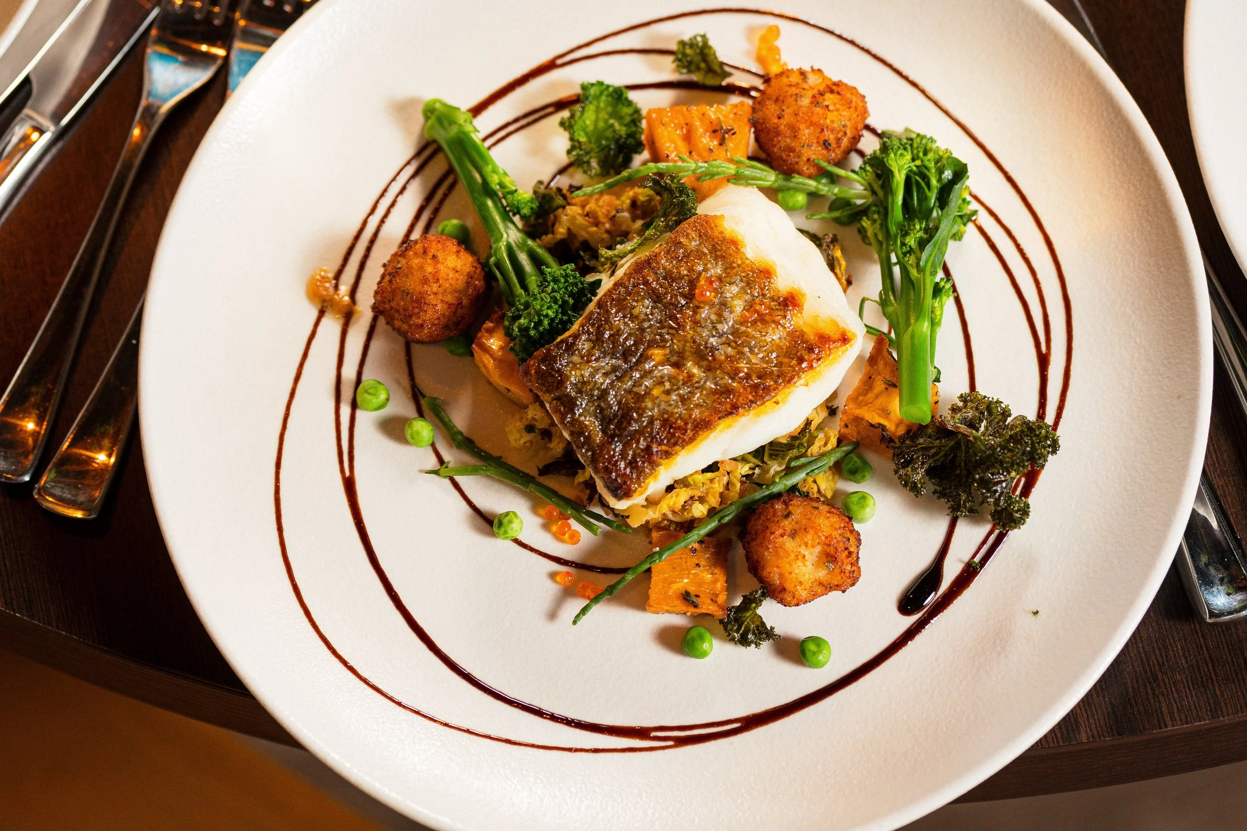 A plated dish with fish, green vegetables, sweet potatoes, and small fried bites, garnished with a decorative swirl of sauce and colorful dots of sauce or garnish.