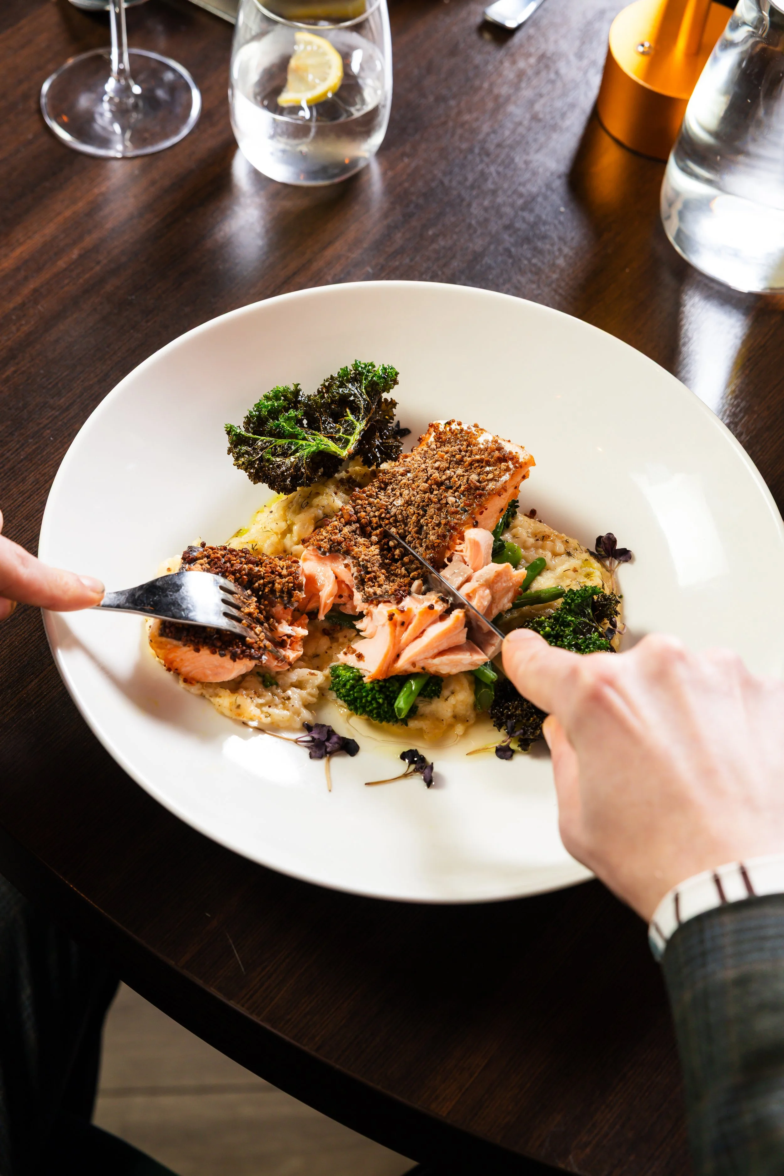 A person is cutting a piece of cooked salmon with a fork and knife on a white plate, which also has greens and mashed potatoes, on a dark wooden table with water glasses and a lit candle nearby.