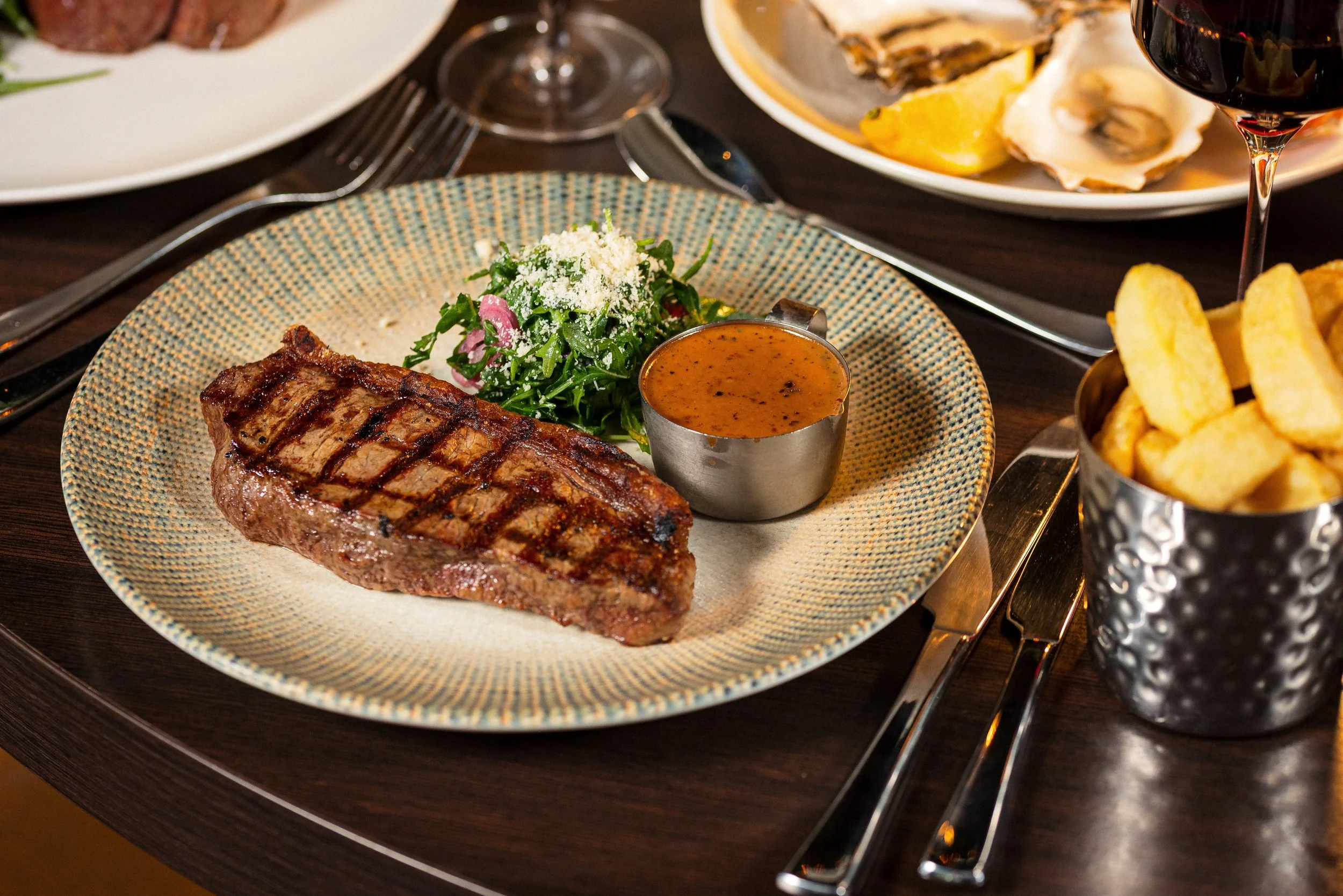 A plate with a grilled steak, a salad with greens and grated cheese, and a small cup of sauce. In the background are other plates with seafood, lemon wedges, and a glass of red wine on a dark table.