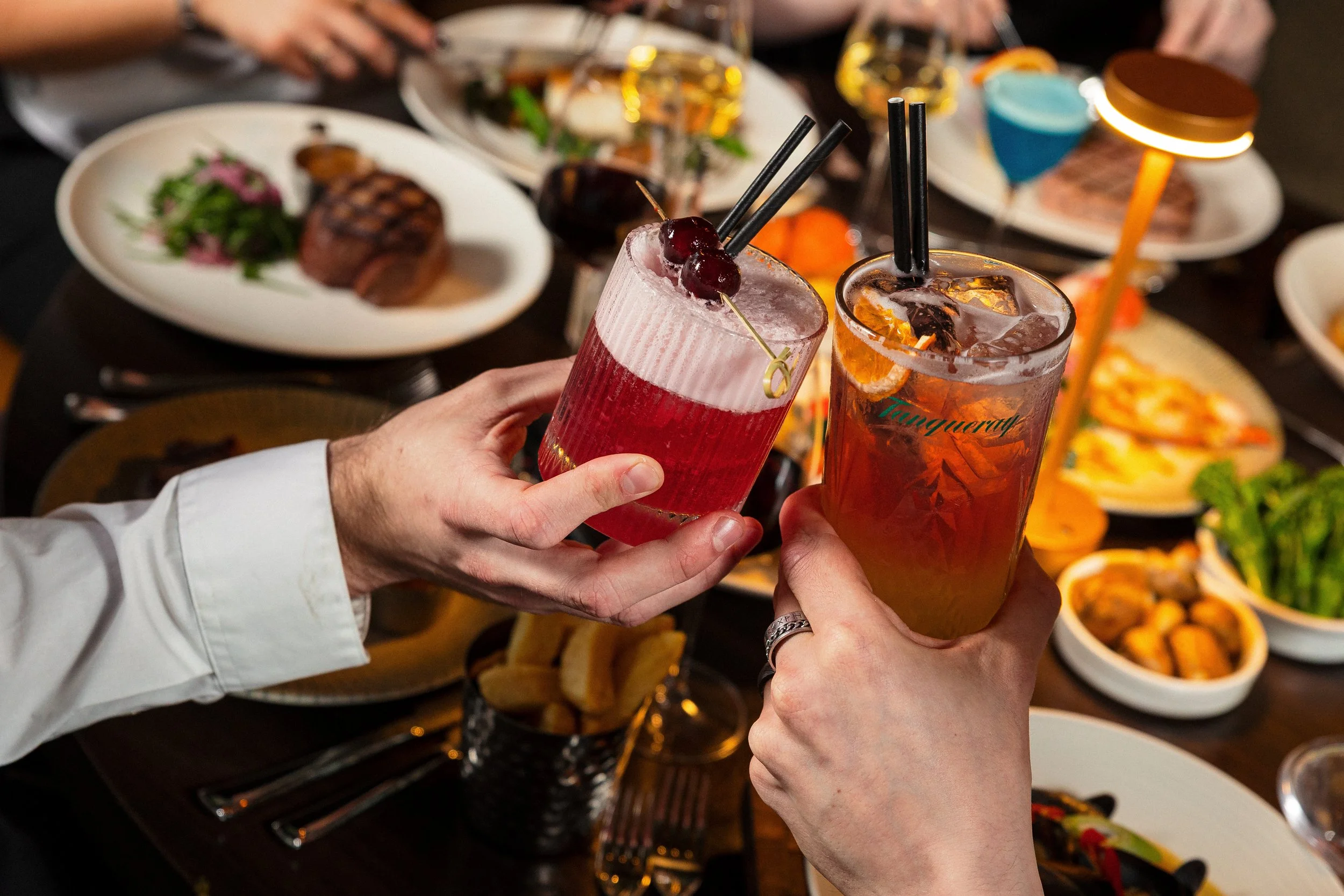 Two people clinking glasses of cocktails at a restaurant or party, with them surrounded by plates of food on a table.