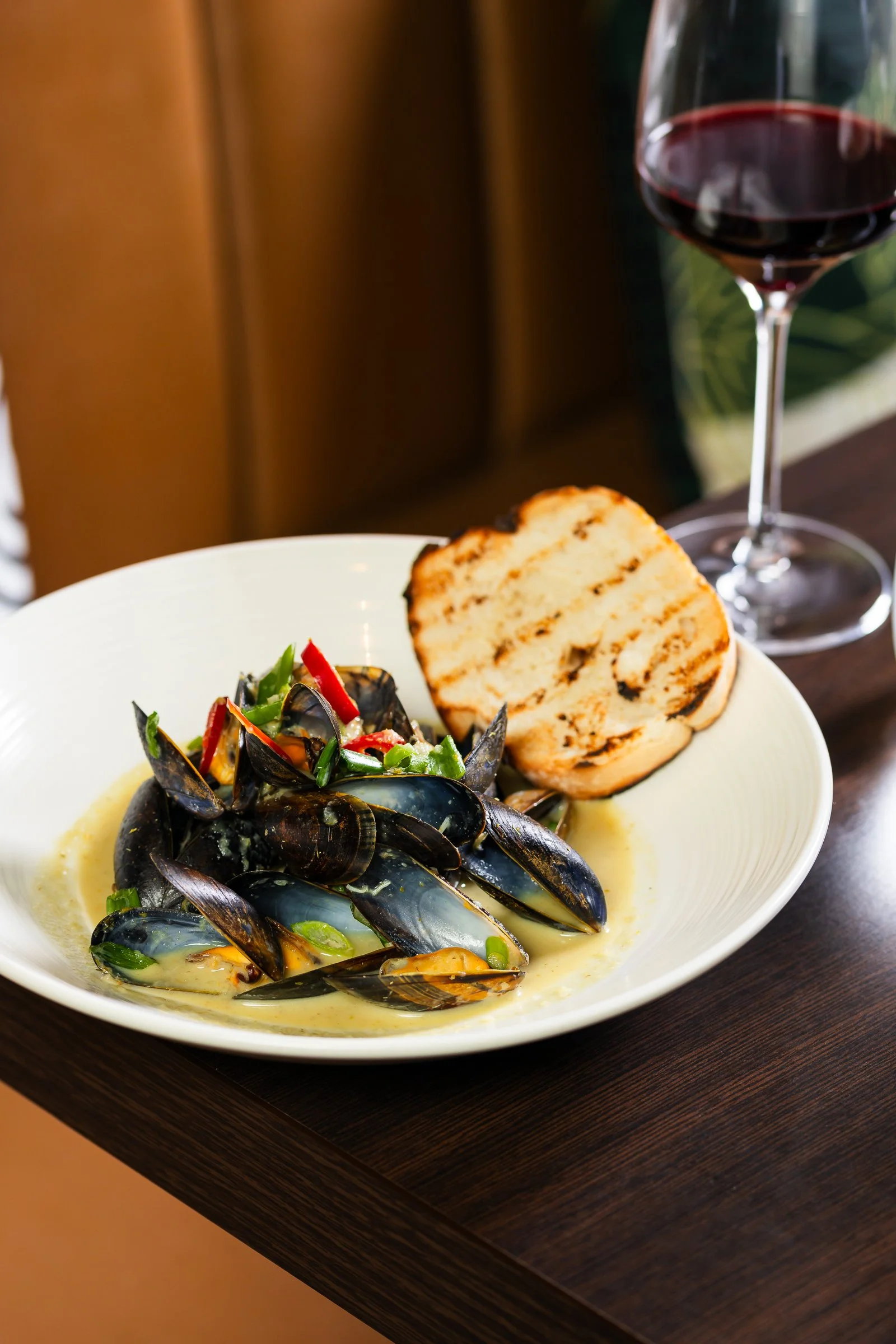 A bowl of mussels with chopped vegetables and herbs, served with a slice of toasted bread, alongside a glass of red wine on a dark wooden table.
