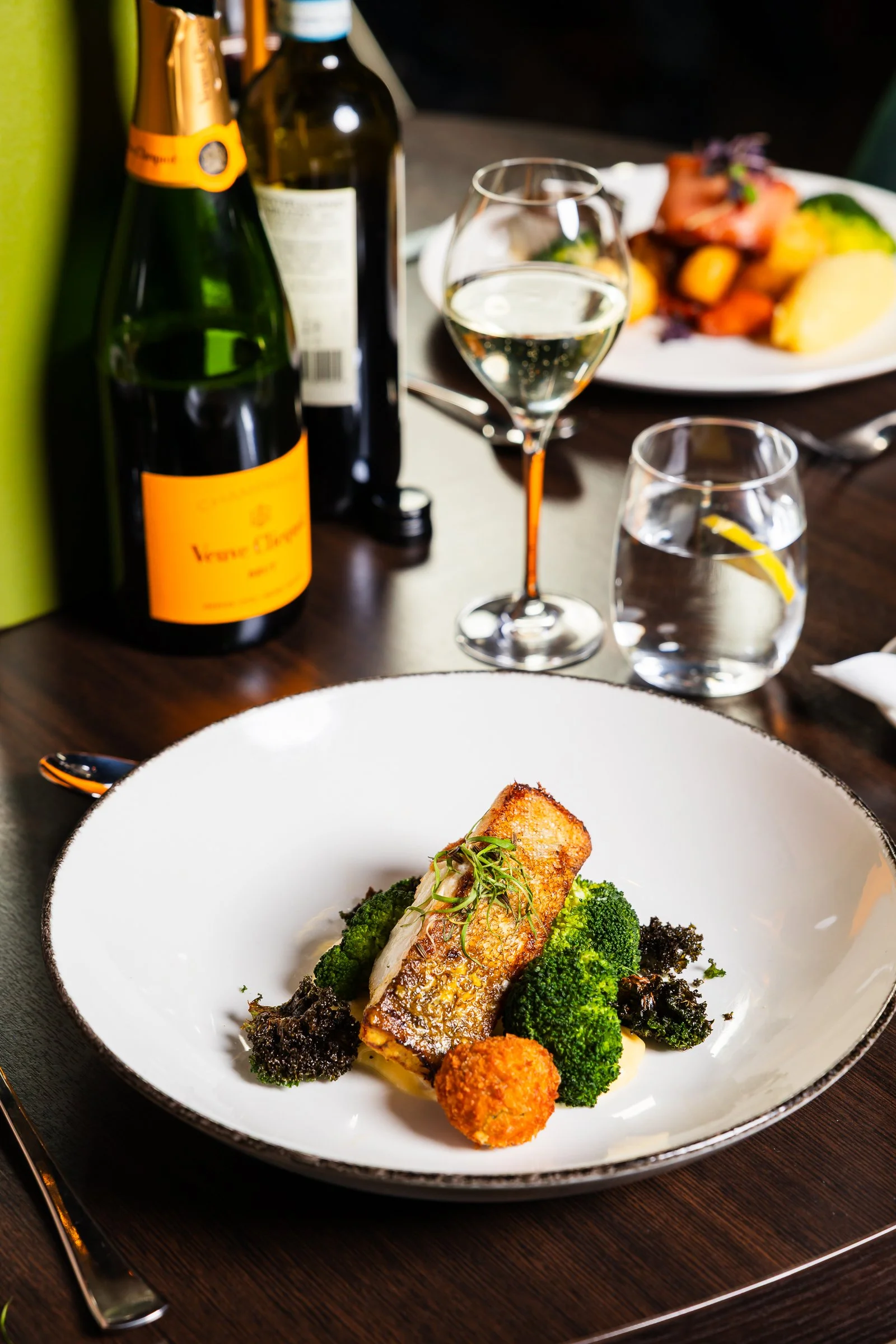 A white plate contains a piece of cooked fish on a bed of broccoli, garnished with microgreens, with two breaded croquettes and orange-colored vegetables on the side. In the background, there are two bottles of sparkling wine and a glass of white win