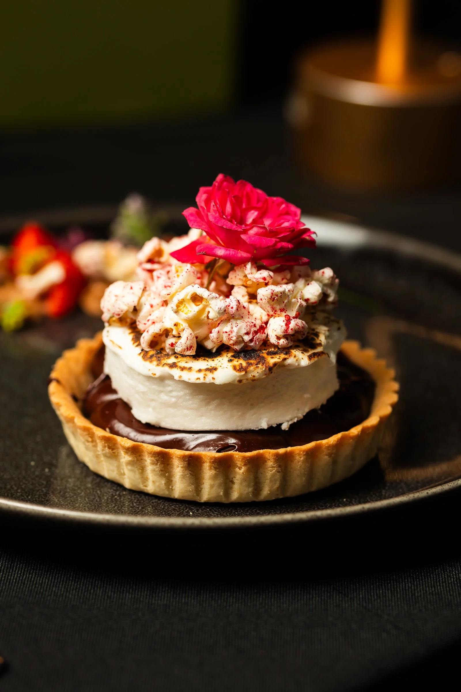 Chocolate tart with whipped cream, pink flower, and strawberries on a black plate