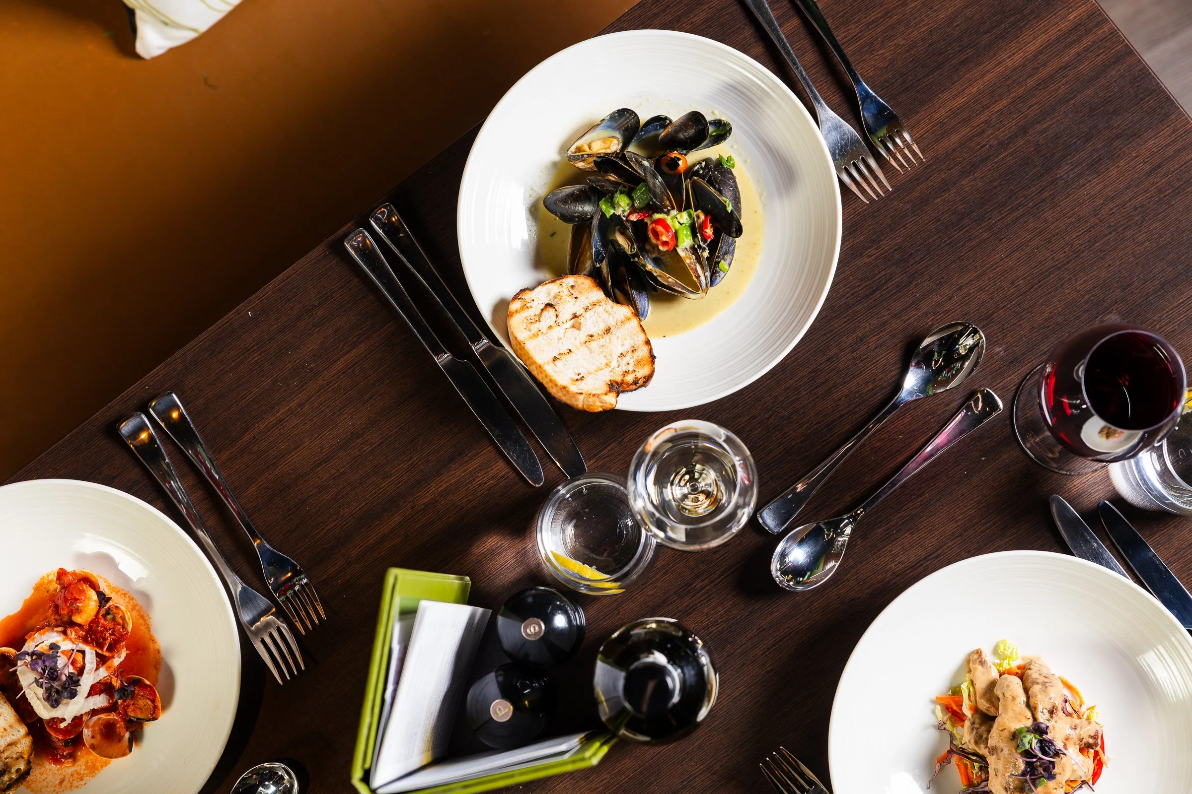 A top-down view of a dinner table with plates of food, including a seafood dish with mussels in a creamy sauce, beside a toasted bread slice. The table is set with forks, knives, spoons, and glasses of red wine and water.