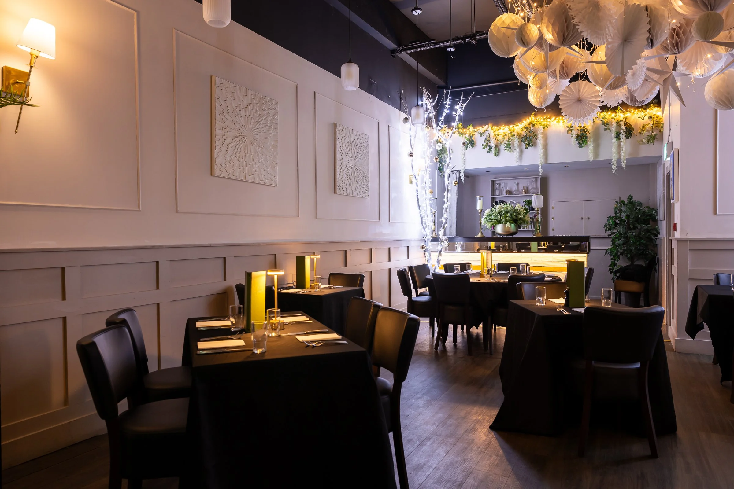 Elegant restaurant interior decorated with hanging paper lanterns, flowers, and string lights, with black tables set for dining.