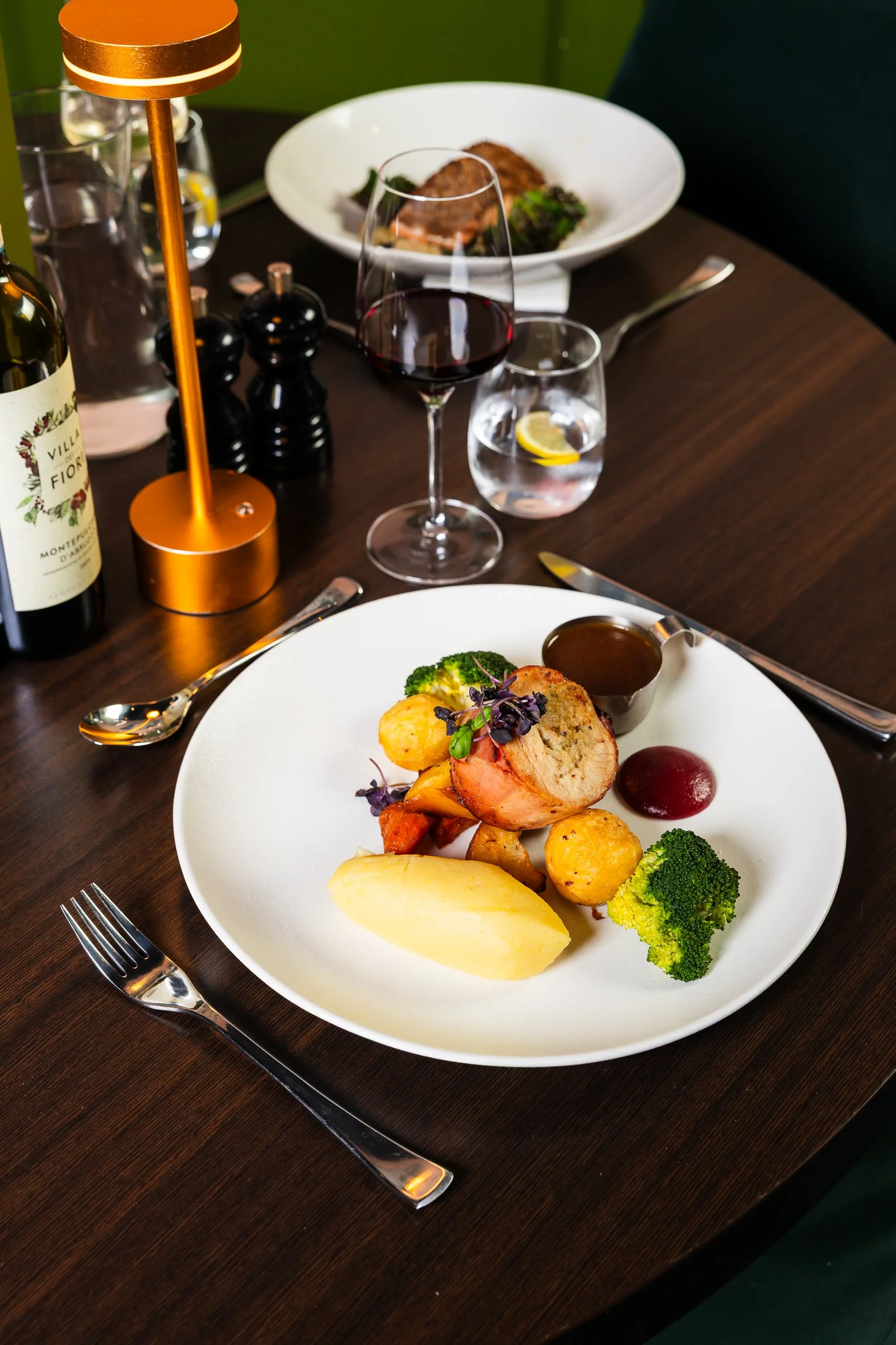 A plate with a roasted chicken breast, broccoli, carrots, cauliflower, purple potatoes, mashed potato, and sauce. In the background, there is a glass of red wine, a glass of water with lemon, a bowl of food, a bottle of red wine, salt and pepper shak