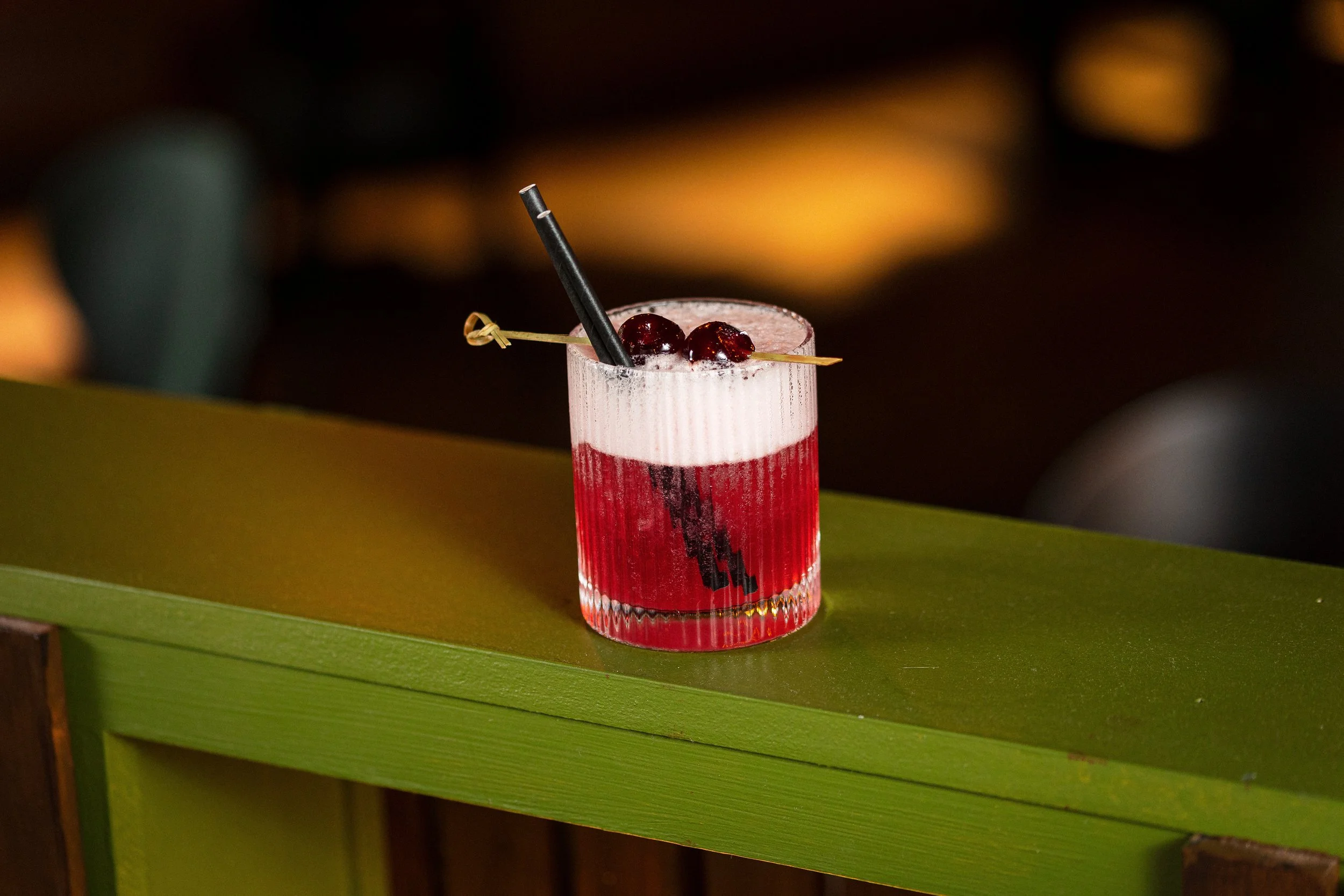 A layered pink and red cocktail garnished with a cherry and served with a black straw and a skewer with two cherries, placed on a green surface at night with blurry warm lights in the background.
