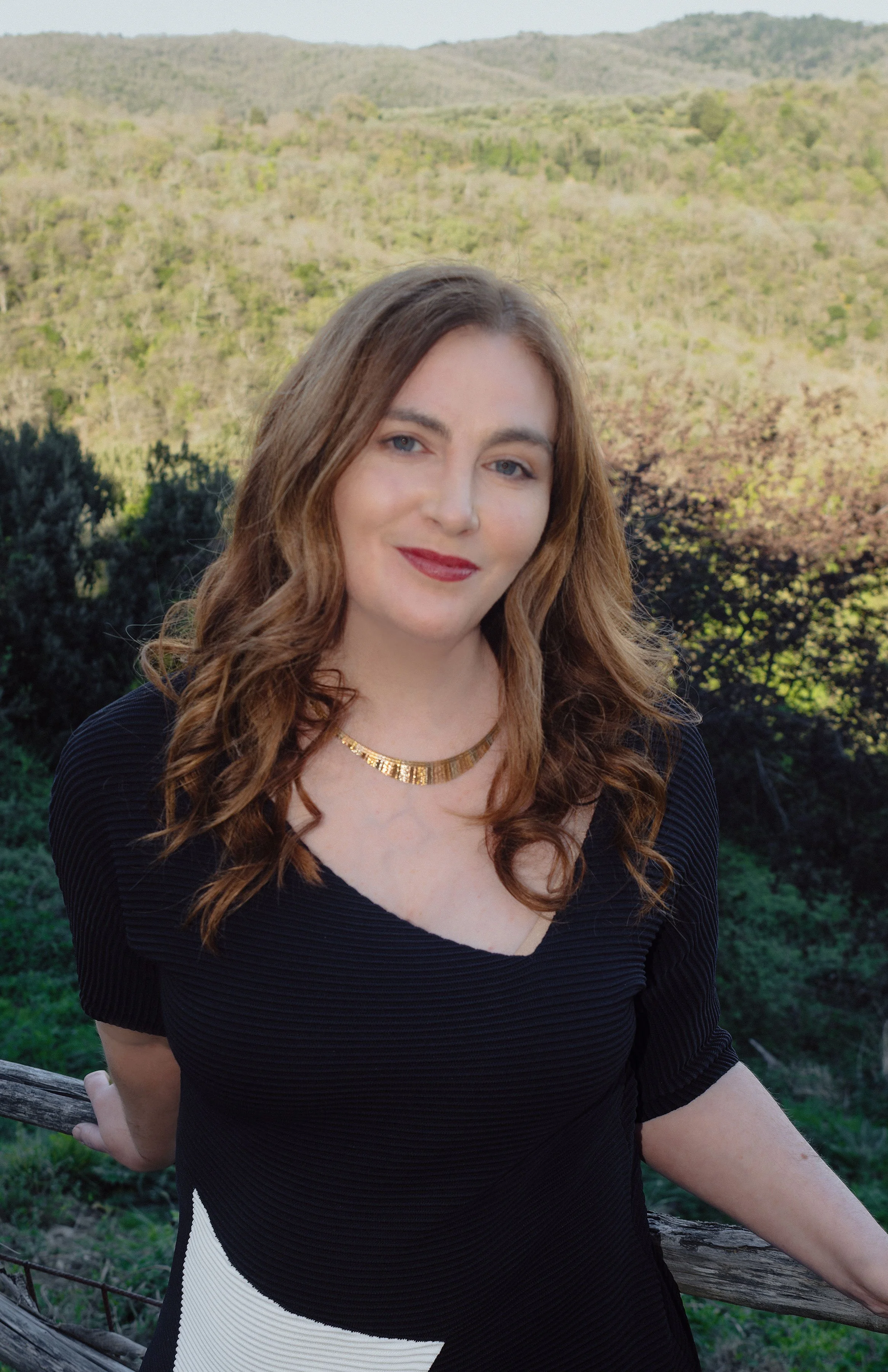 Rachel Emerson with long wavy red hair, wearing a black top with cream-colored geometric patterns and a gold necklace, is standing outdoors near a wooden fence with green hills and trees in the background.