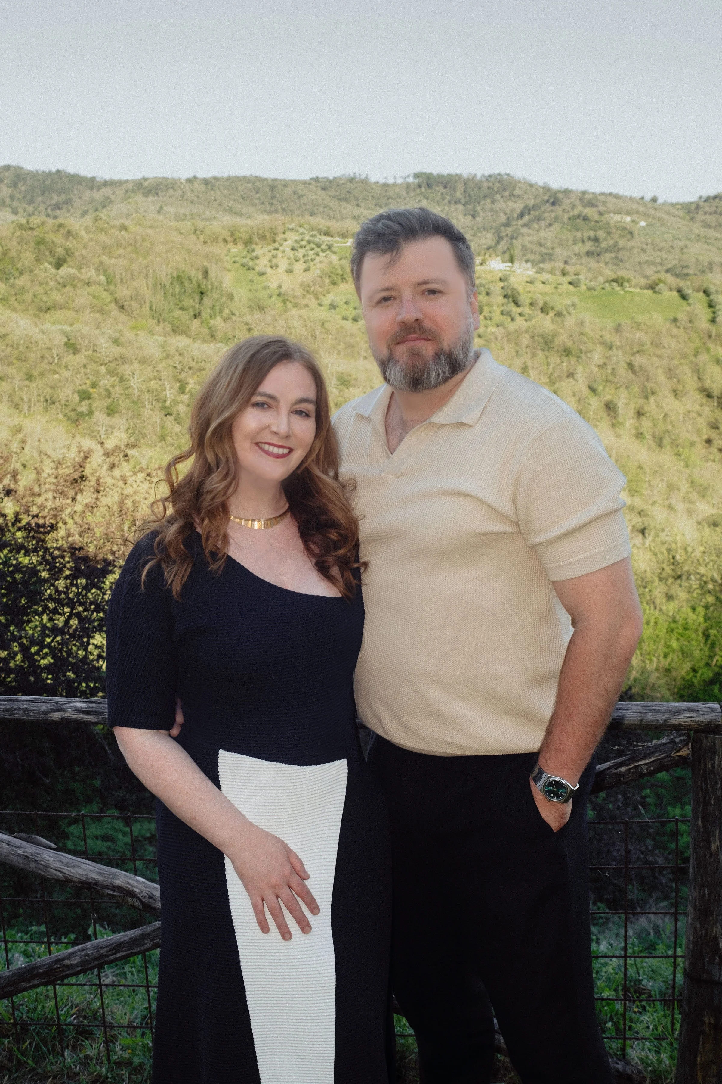A man and woman stand outdoors in front of a green hillside, smiling and posing for a photo. The woman has long, wavy brown hair and is wearing a black top with a gold necklace, while the man has short dark hair and a beard, wearing a light-colored polo shirt with black pants and a watch on his left wrist.