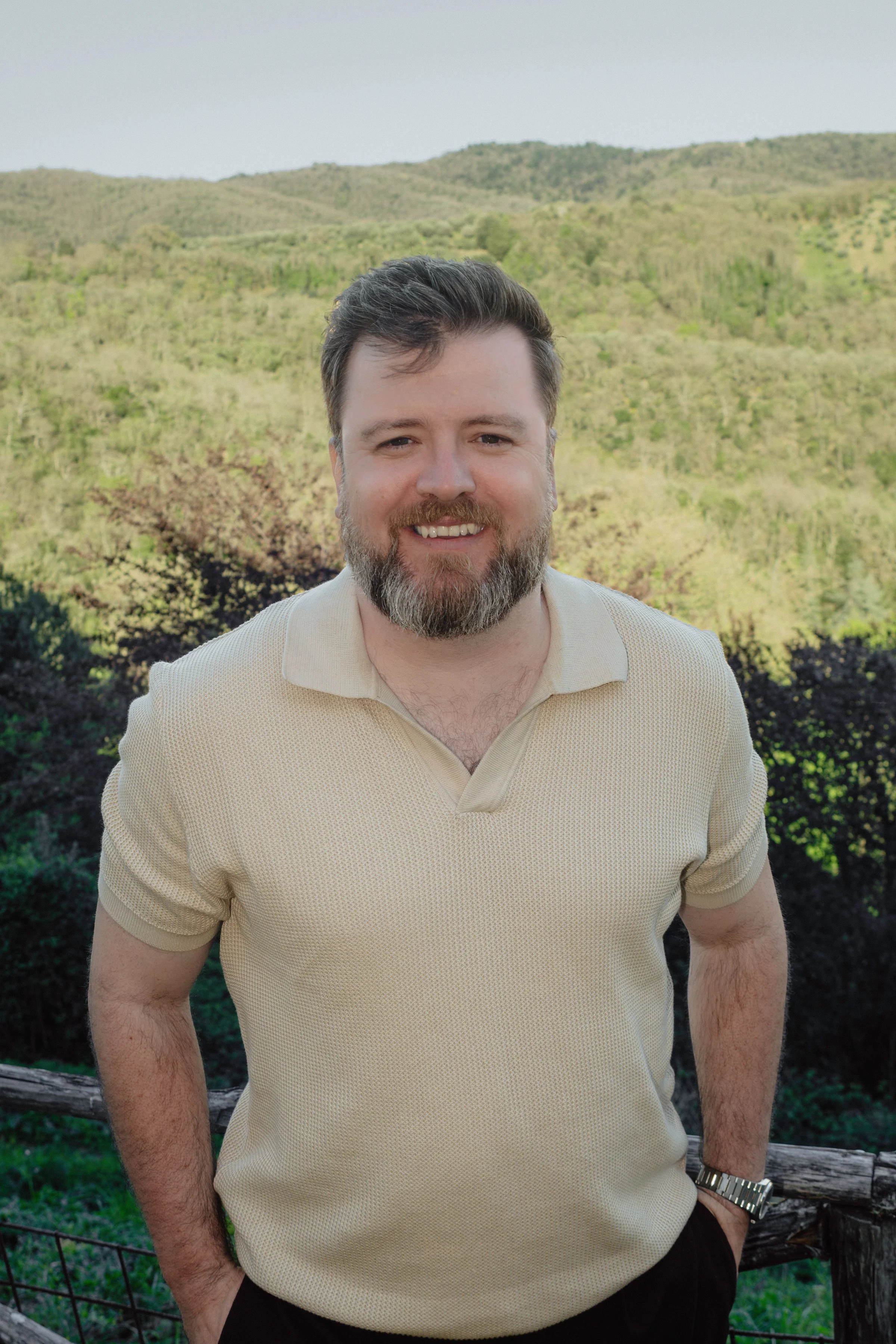 Portrait of a smiling Ian Sumpter with short dark hair and a beard, wearing a beige polo shirt and a watch, standing outdoors with a mountain and forested landscape in the background.